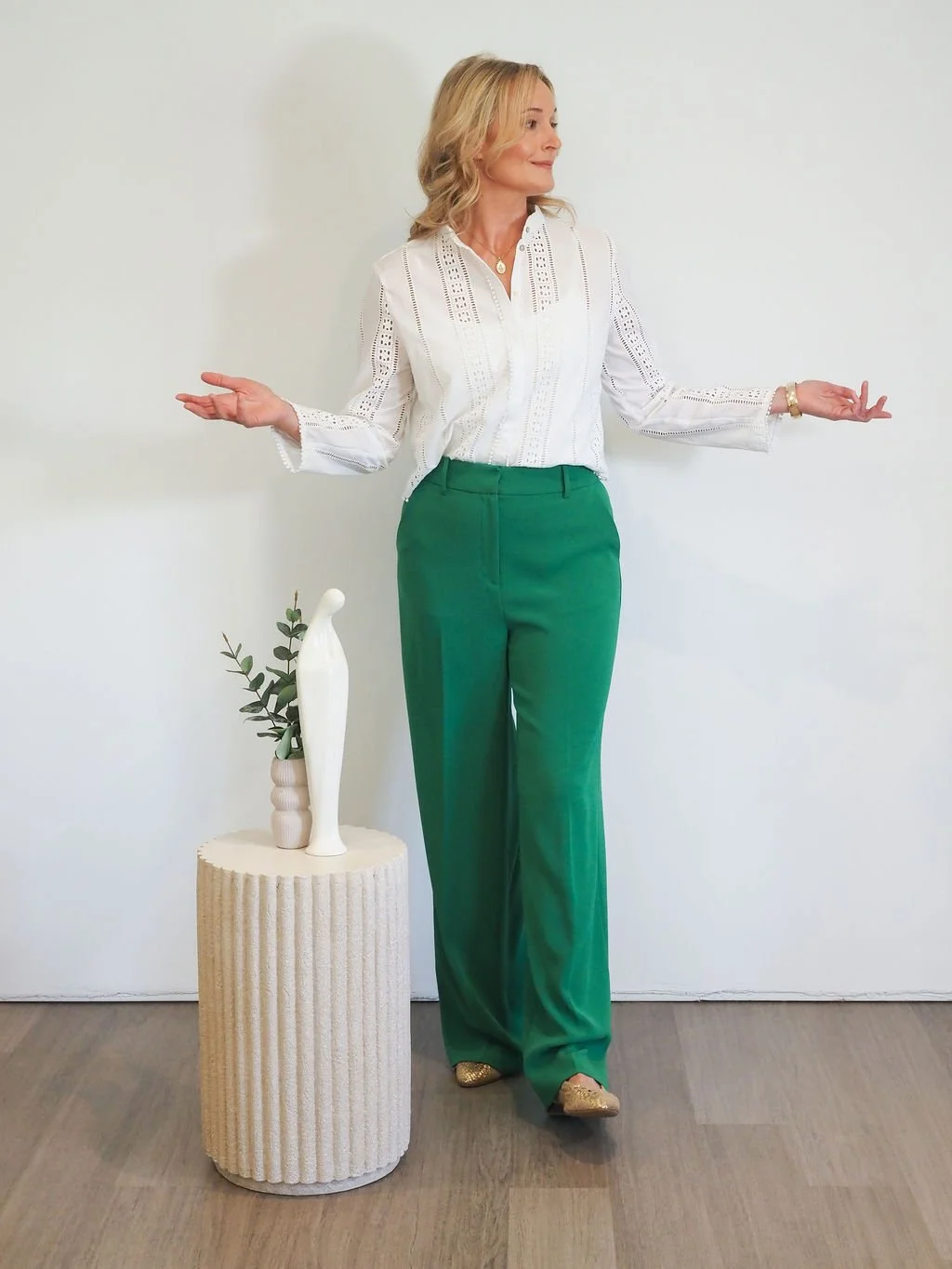 A woman with blonde hair in a white blouse and green pants standing against a plain white wall, with her arms outstretched to her sides and a decorative vase on a small cylindrical table to her left.