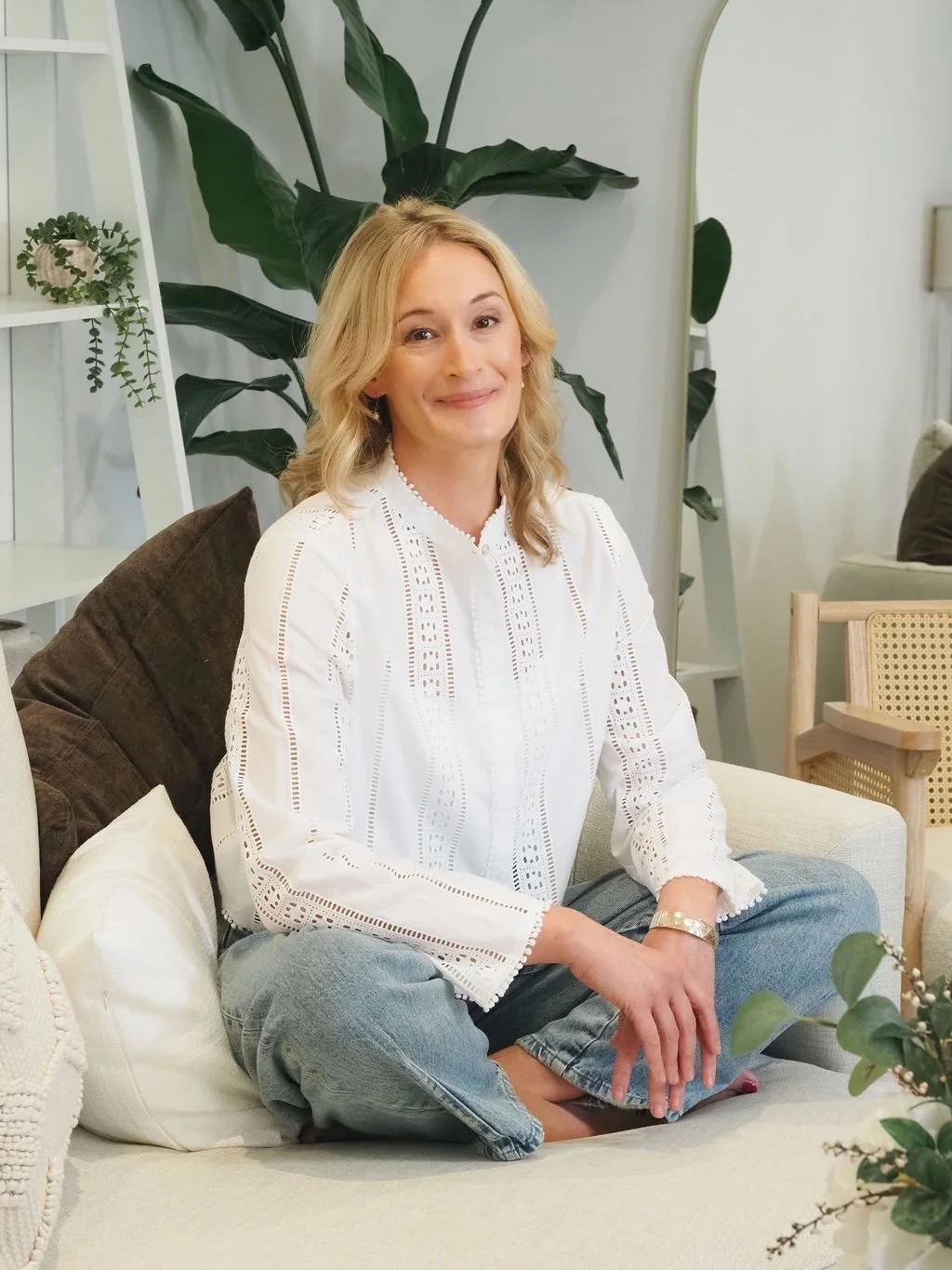 A woman with blonde hair sitting cross-legged on a sofa in a cozy, well-decorated living room with plants and pillows.