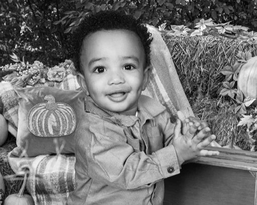 Smiling young child sitting outdoors near plants, with a decorated pillow featuring a pumpkin design.