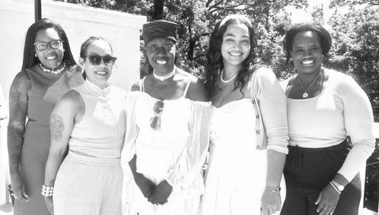 Five women smiling and standing together outdoors in sunlight, dressed in light-colored clothing.