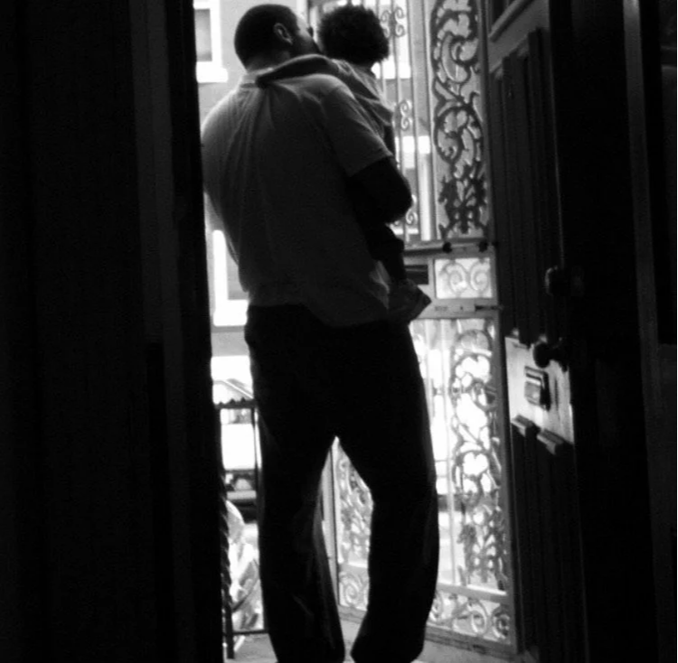 A man holding a child standing at an open door with a decorative balcony railing in the background in black and white.