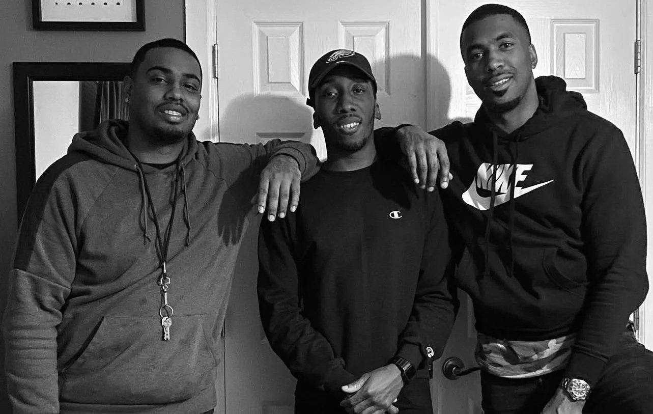Three young men standing together indoors, smiling at the camera, with their arms around each other's shoulders.