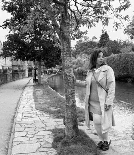 Woman standing by a tree on a sidewalk near a canal or river, wearing a coat and dress.