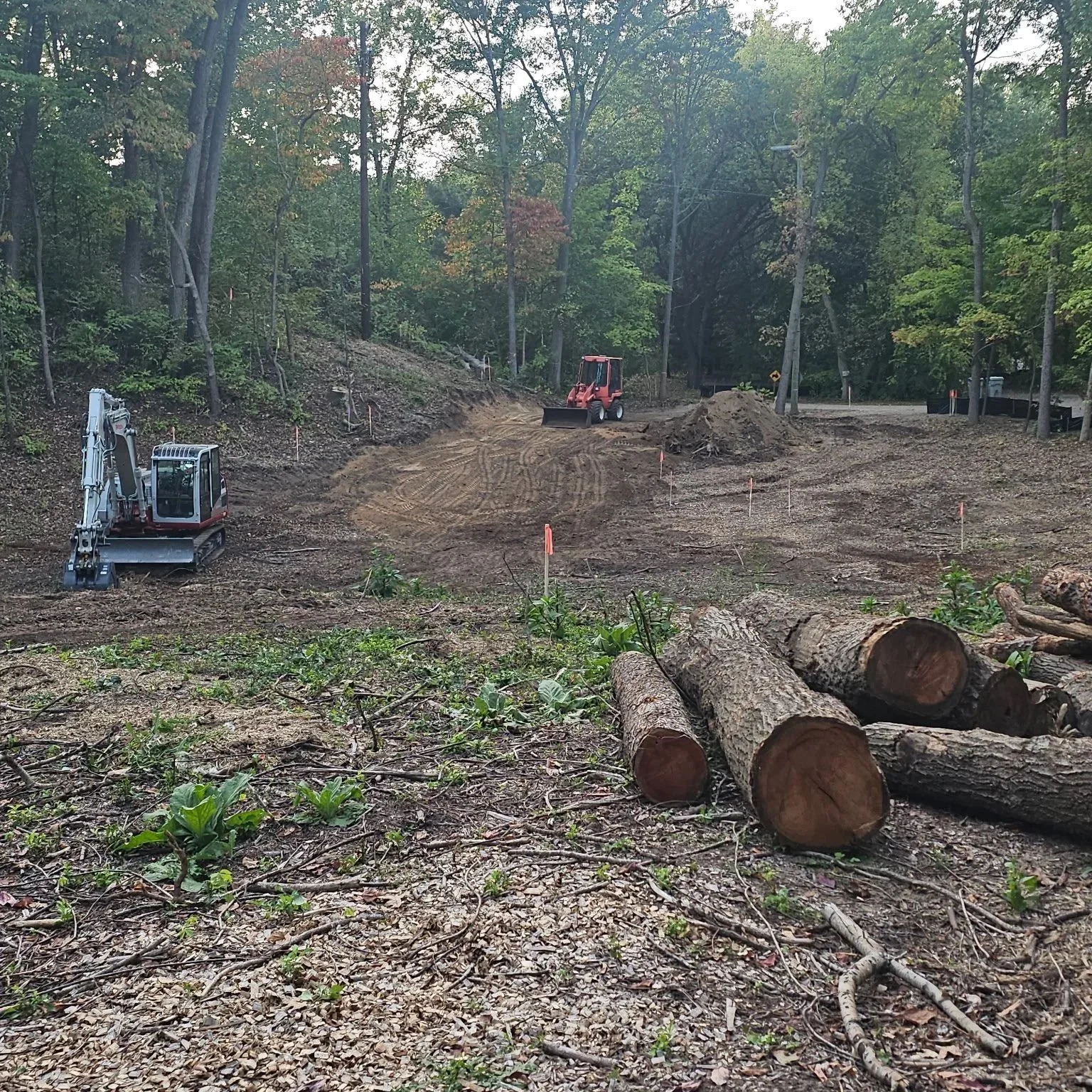 Construction site with trees, a dirt road, construction machinery including a compact excavator and a bulldozer, and logs in the foreground.