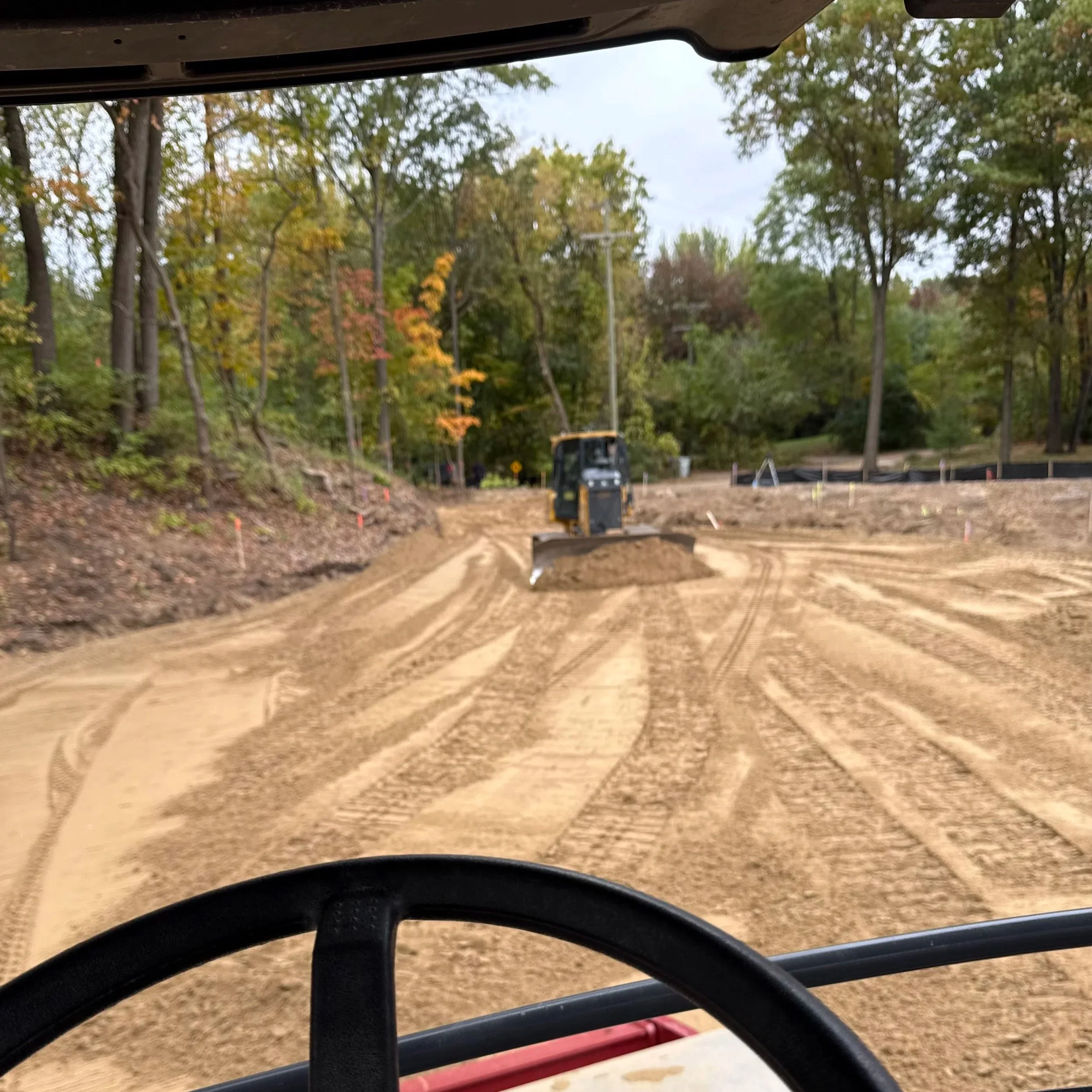 View from inside a construction vehicle showing a dirt road being graded by a road roller, with trees and construction markers on a cloudy day.
