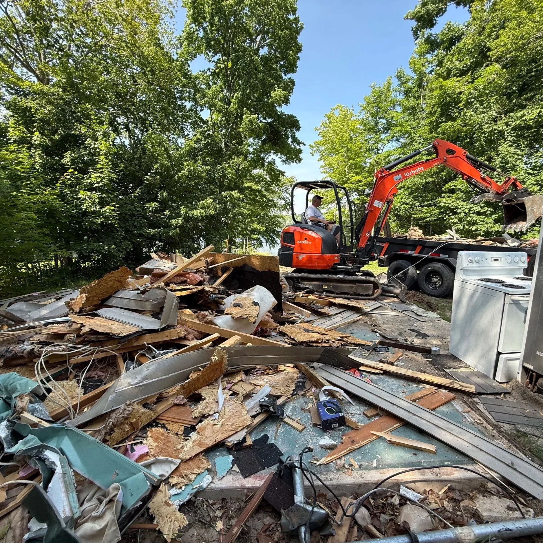 A person using a small orange excavator to clear debris in a backyard with trees, appliances, and construction materials.