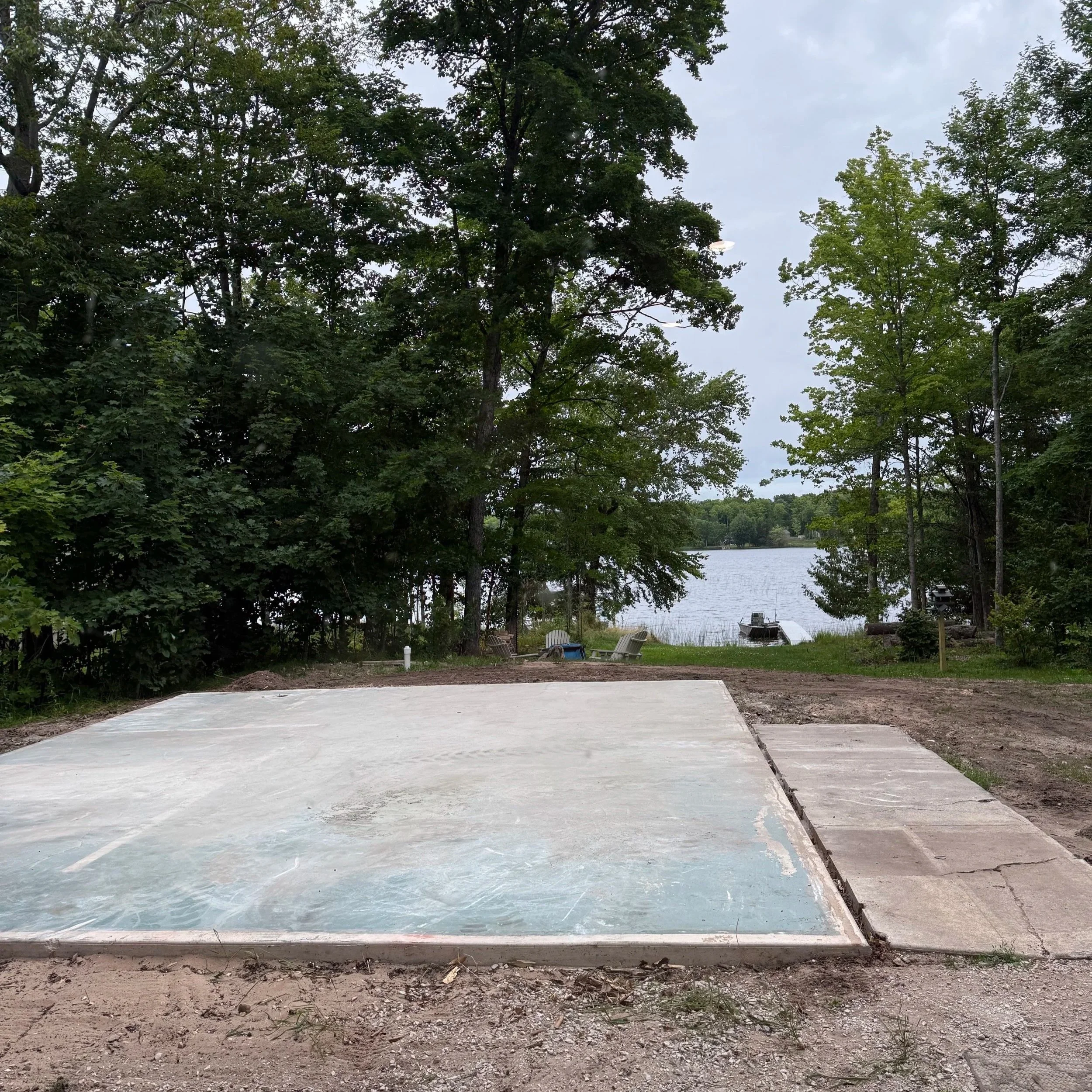 Empty concrete pad near a lake, with trees and outdoor chairs in the background.