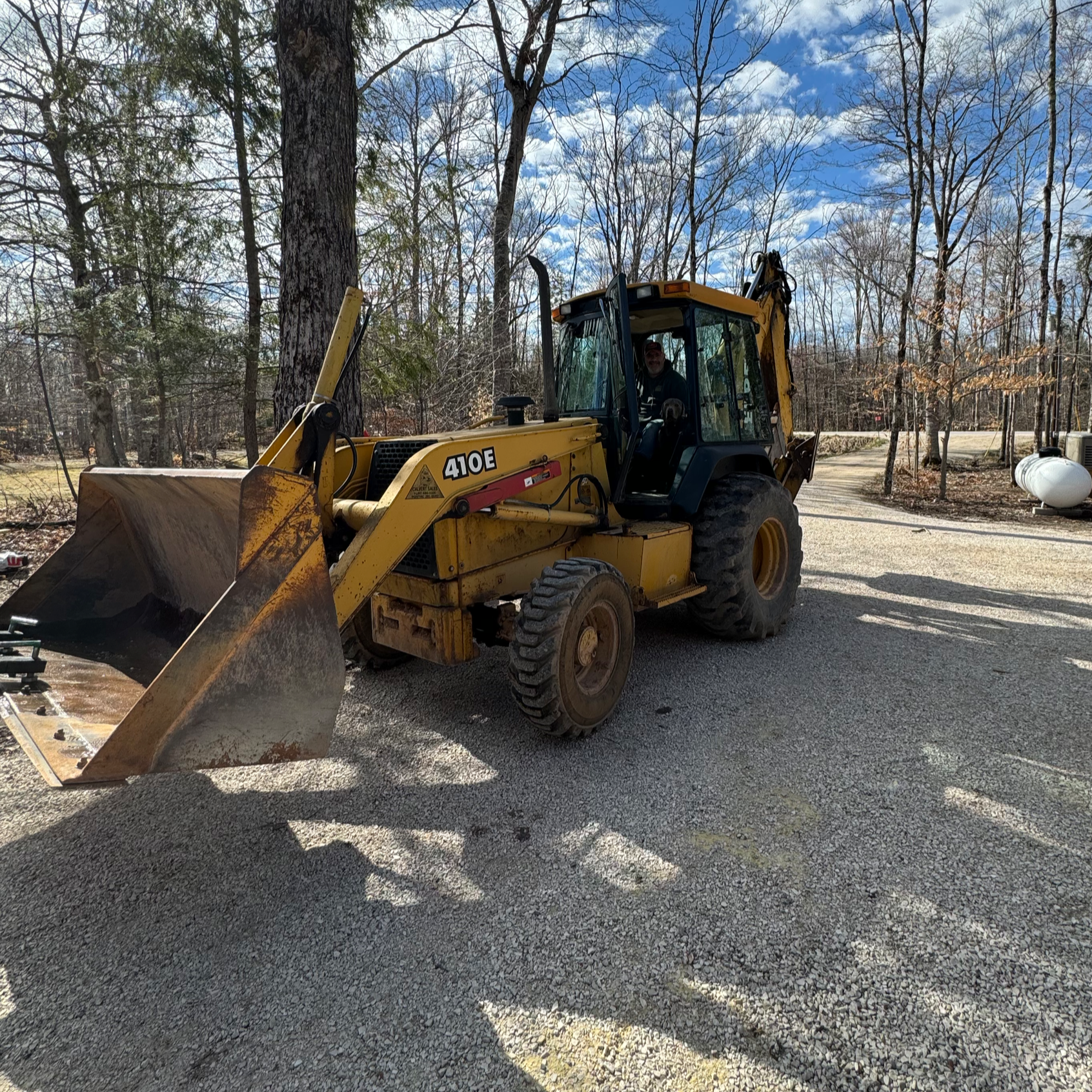 A man driving a yellow bulldozer labeled 410E on a gravel road, with trees and a propane tank in the background under a partly cloudy sky.