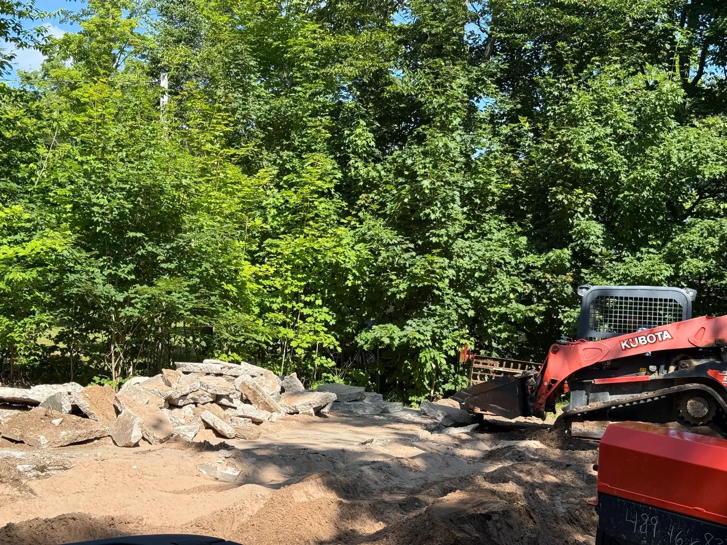 Construction site with a small Kubota excavator and piles of rocks, set against a backdrop of leafy green trees under a clear blue sky.
