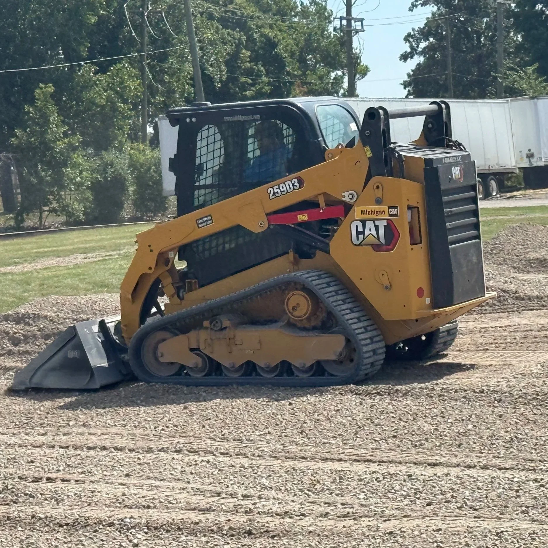 A yellow Caterpillar skid steer loader with tracks, working on a dirt surface at a construction site, with a trailer and trees in the background.