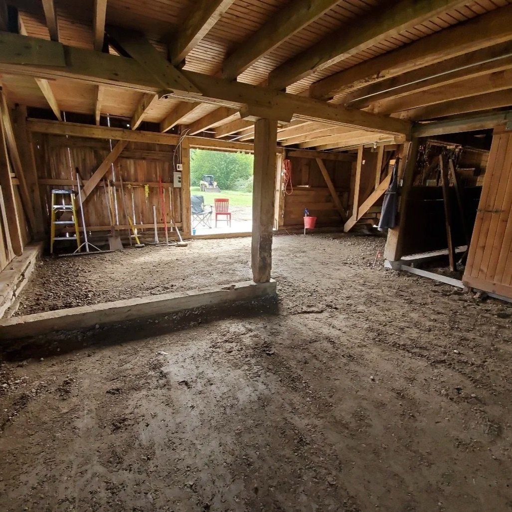 Inside a wooden barn with a dirt floor, construction tools and equipment are visible along the walls. Outside, a grassy area with chairs, a tractor, and a person is seen through the open door.