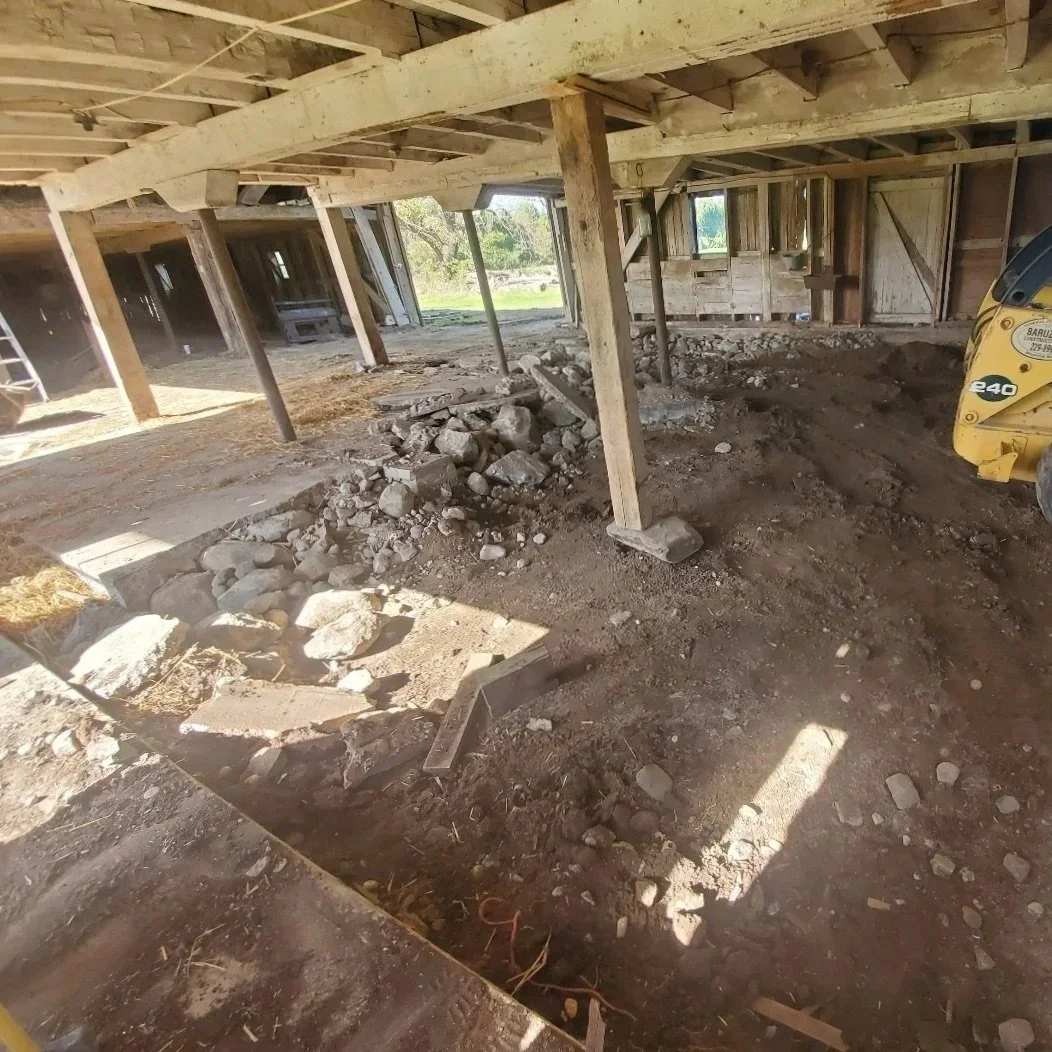 Inside a building under renovation with exposed wooden beams, support poles, dirt floor, scattered rocks, and construction equipment.