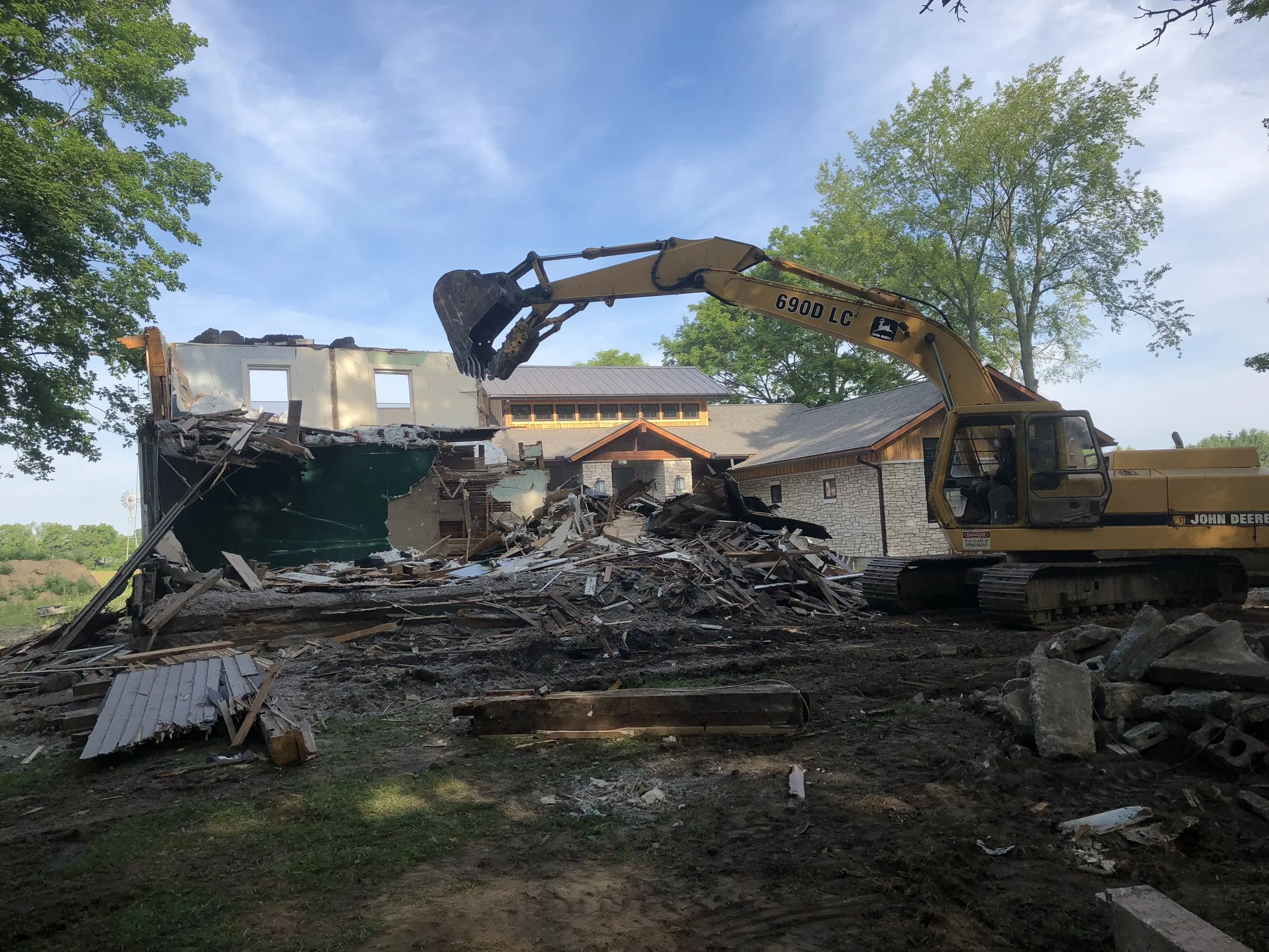 Excavator demolishing a building with debris scattered on the ground and a house in the background.