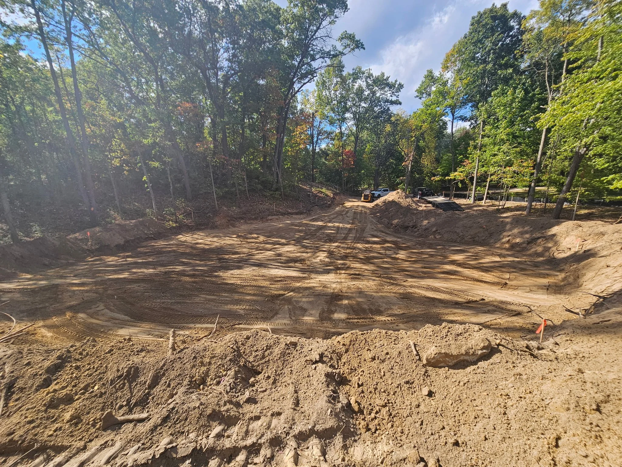 Construction site in a wooded area with cleared dirt, construction vehicles, and parked cars in the background.