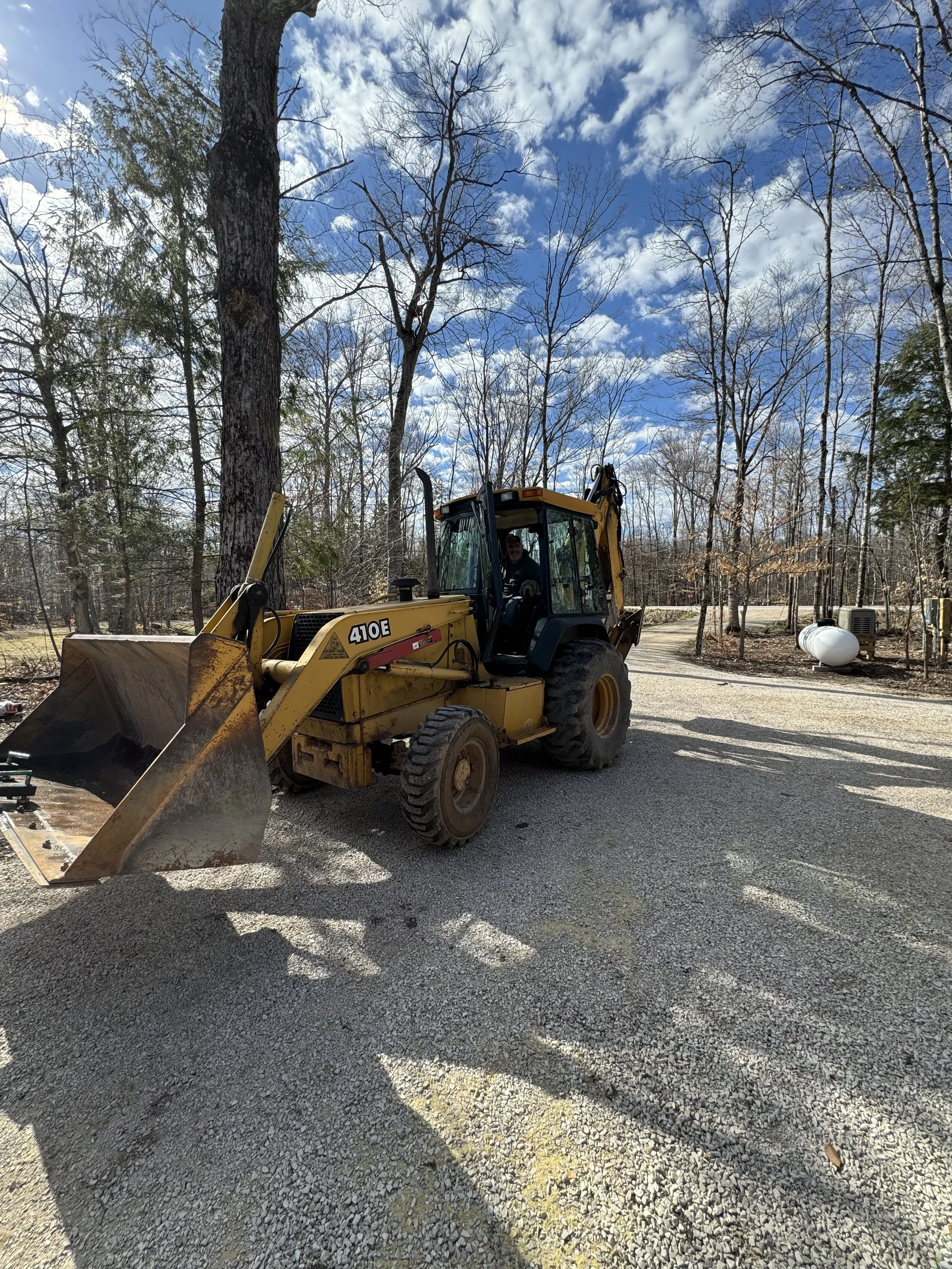 Yellow backhoe loader tractor on gravel driveway in wooded area on a sunny day with blue sky and clouds, man inside the cab.