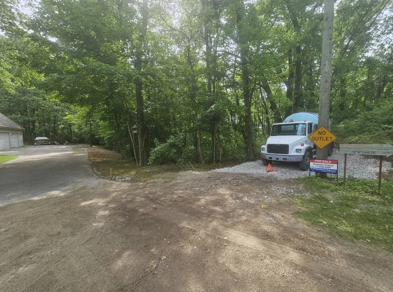 A dirt driveway with a white truck parked beside a tree, sign that reads 'NO OUTLET,' and a 'FOR SALE' sign. Dense greenery and trees surround the area.