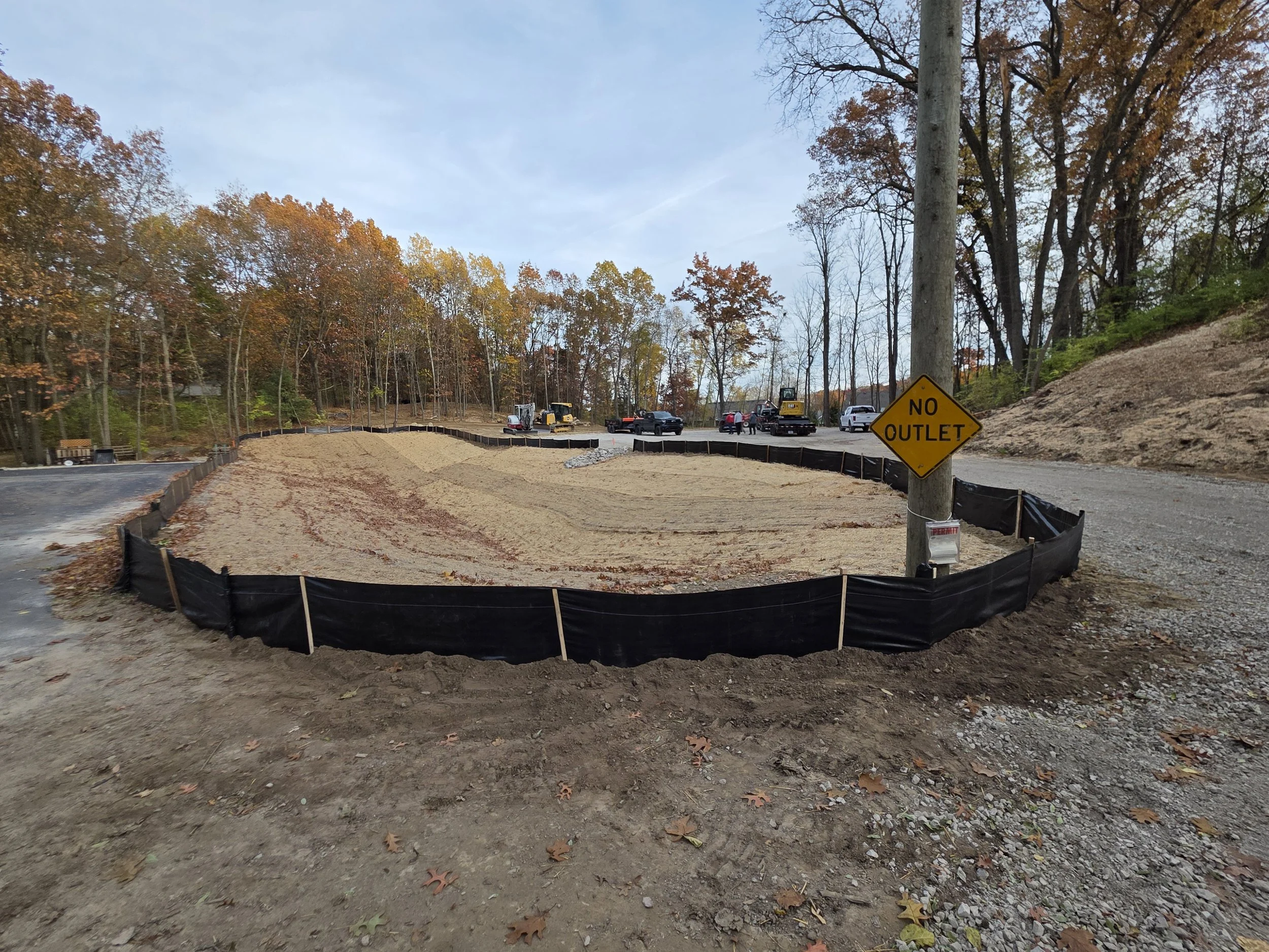 A construction site with a sand-filled area surrounded by black barriers and a yellow 'No Outlet' sign, with a forested background and parked construction vehicles.