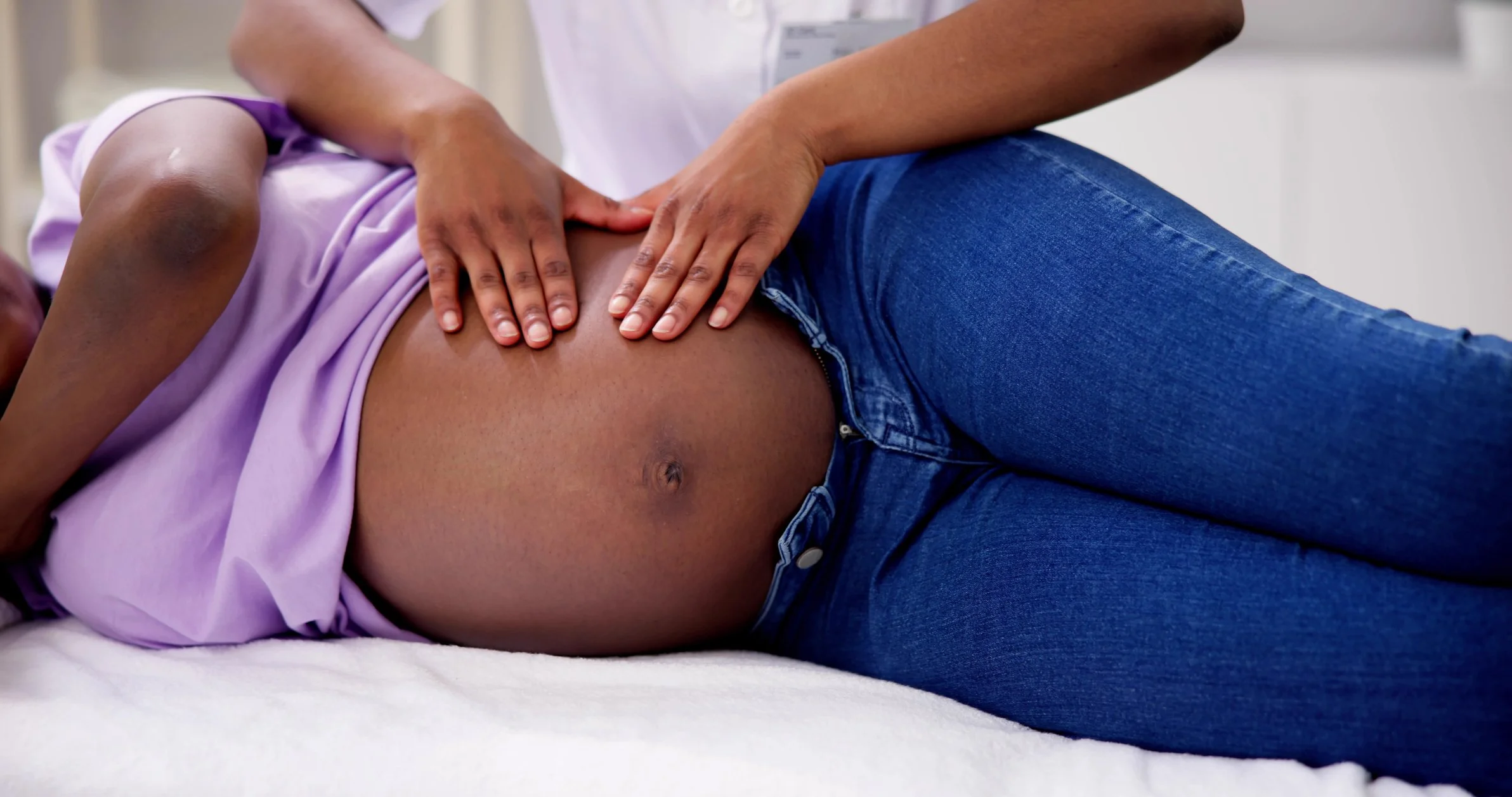 A healthcare professional performs a manual examination or treatment on a patient's abdomen, who is lying on their side wearing a purple top and blue jeans.