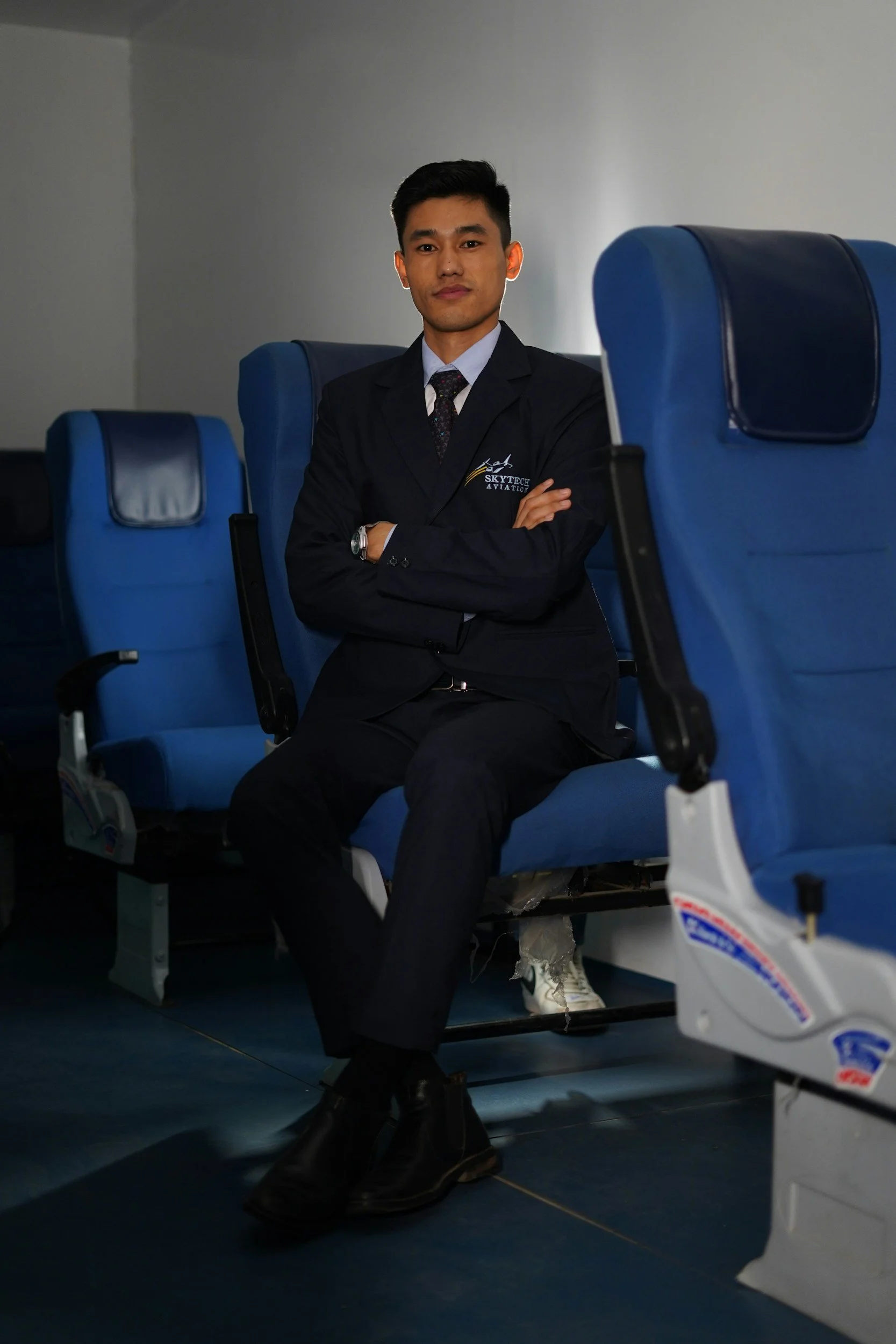 A young man in a dark suit and tie sitting in an airport or train station with blue seats, arms crossed and looking at the camera.