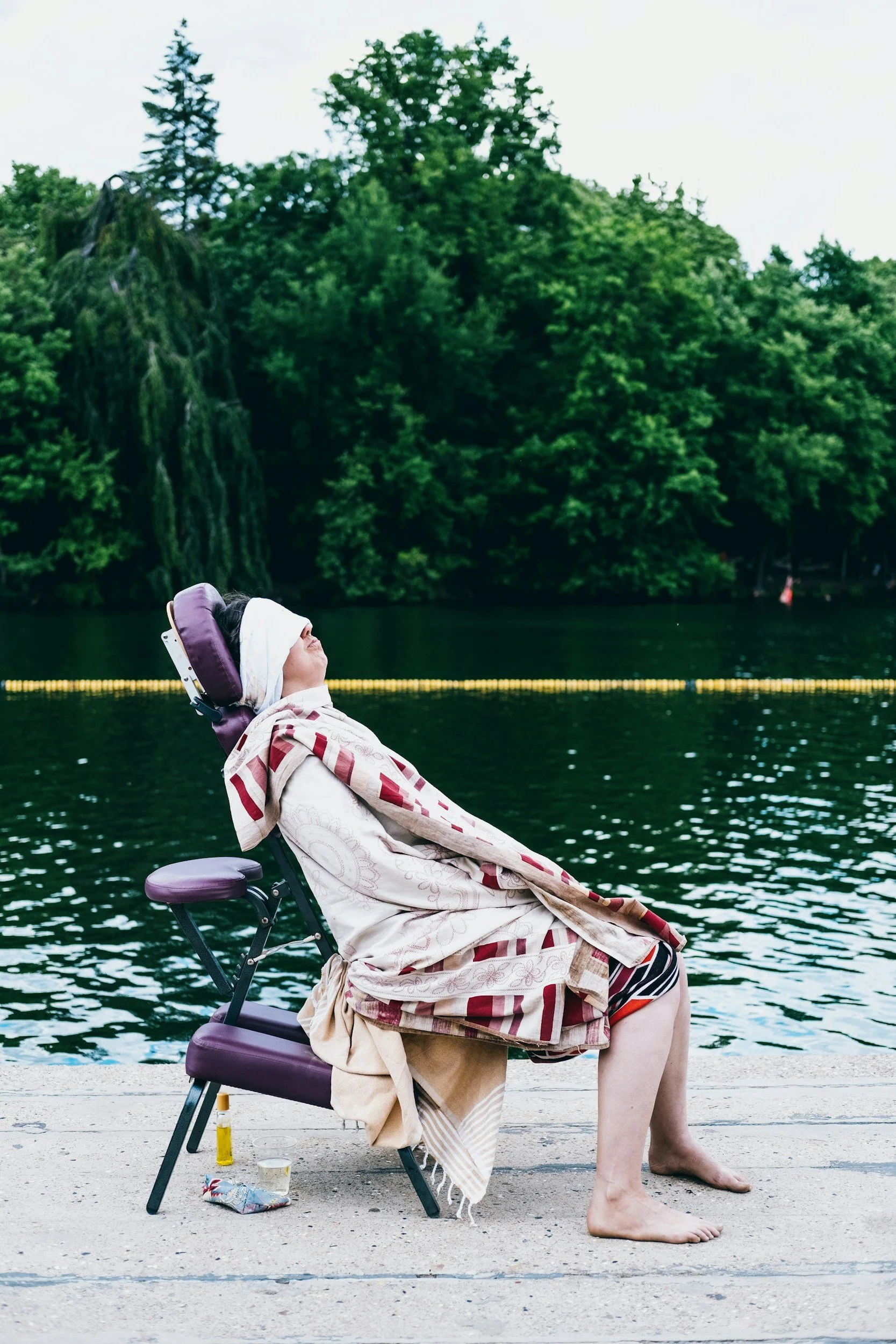 Person sitting in a wheelchair outdoors by a body of water, with a lush green tree line and cloudy sky in the background. The person is covered with a blanket, wearing a headscarf, and has various personal items on the ground nearby.