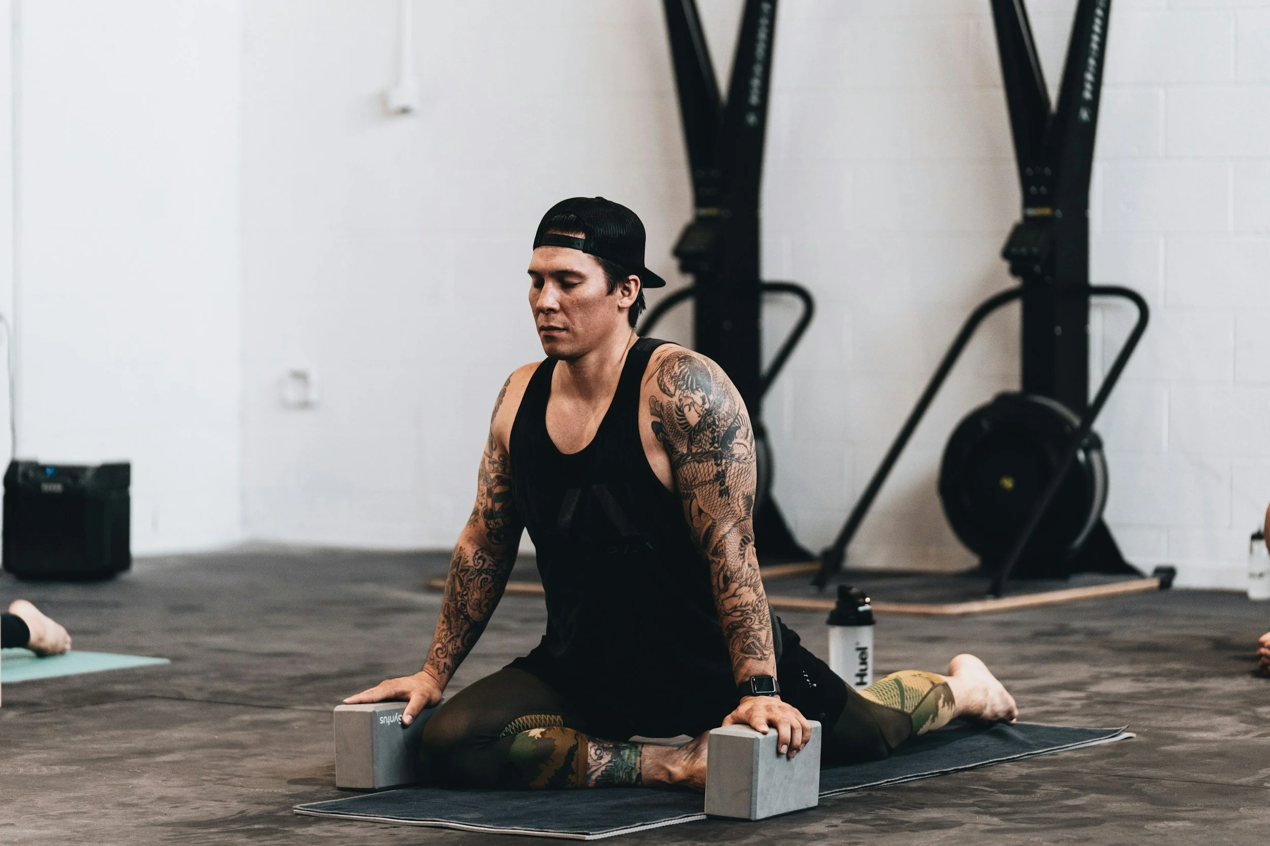A woman with tattoos on her arms practicing yoga in a gym, holding a yoga pose with her hands on blocks on the floor, wearing a black tank top, leggings, and a backwards baseball cap.