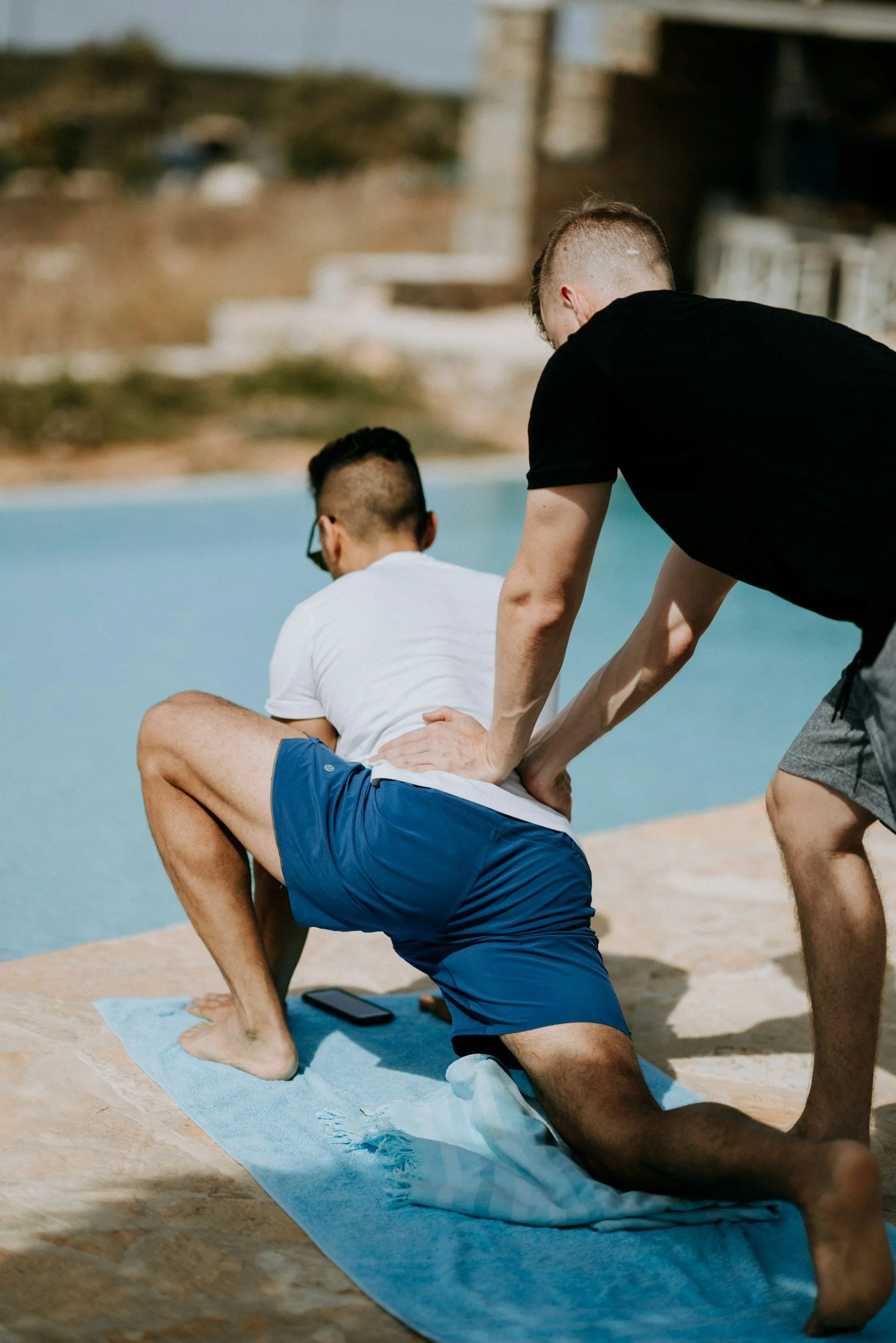 Two men practicing yoga outdoors on a blue mat by the water, one assisting the other with a forward bend pose.