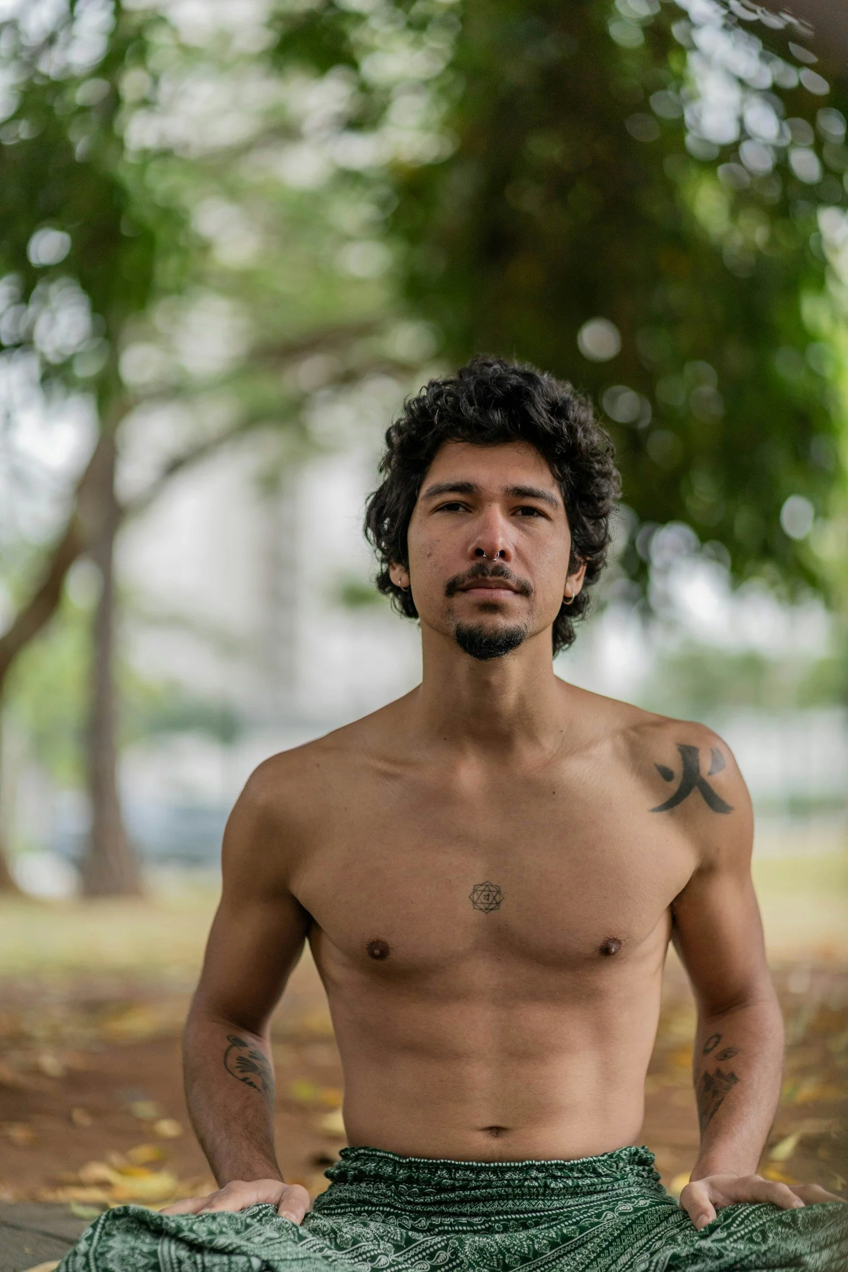 A shirtless man with tattoos on his chest and arms, sitting outdoors on a yoga mat in a park with green trees and leaves in the background.