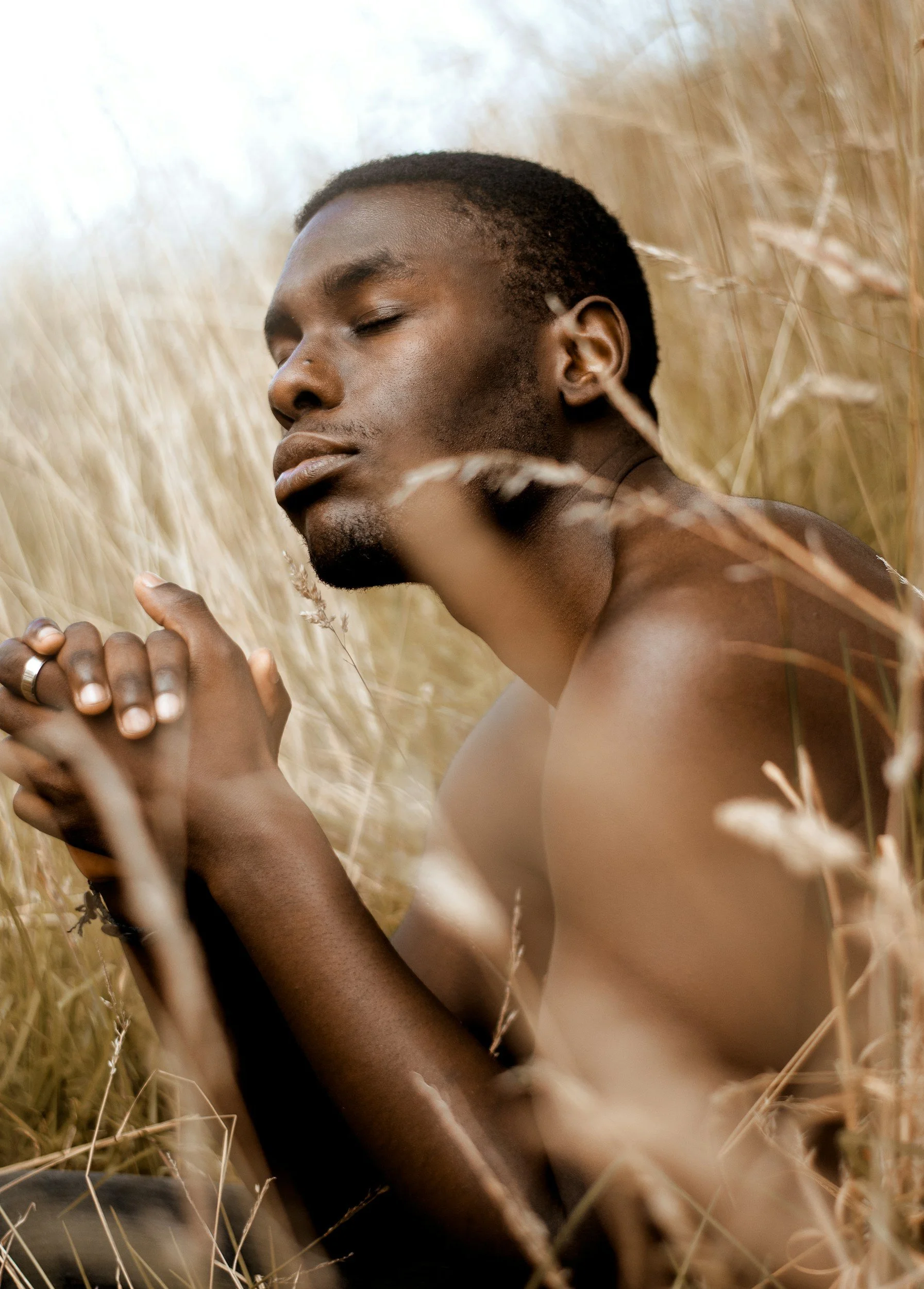 A young man with dark skin and short hair is lying in a field of tall, dry grass with his eyes closed, appearing peaceful and relaxed.