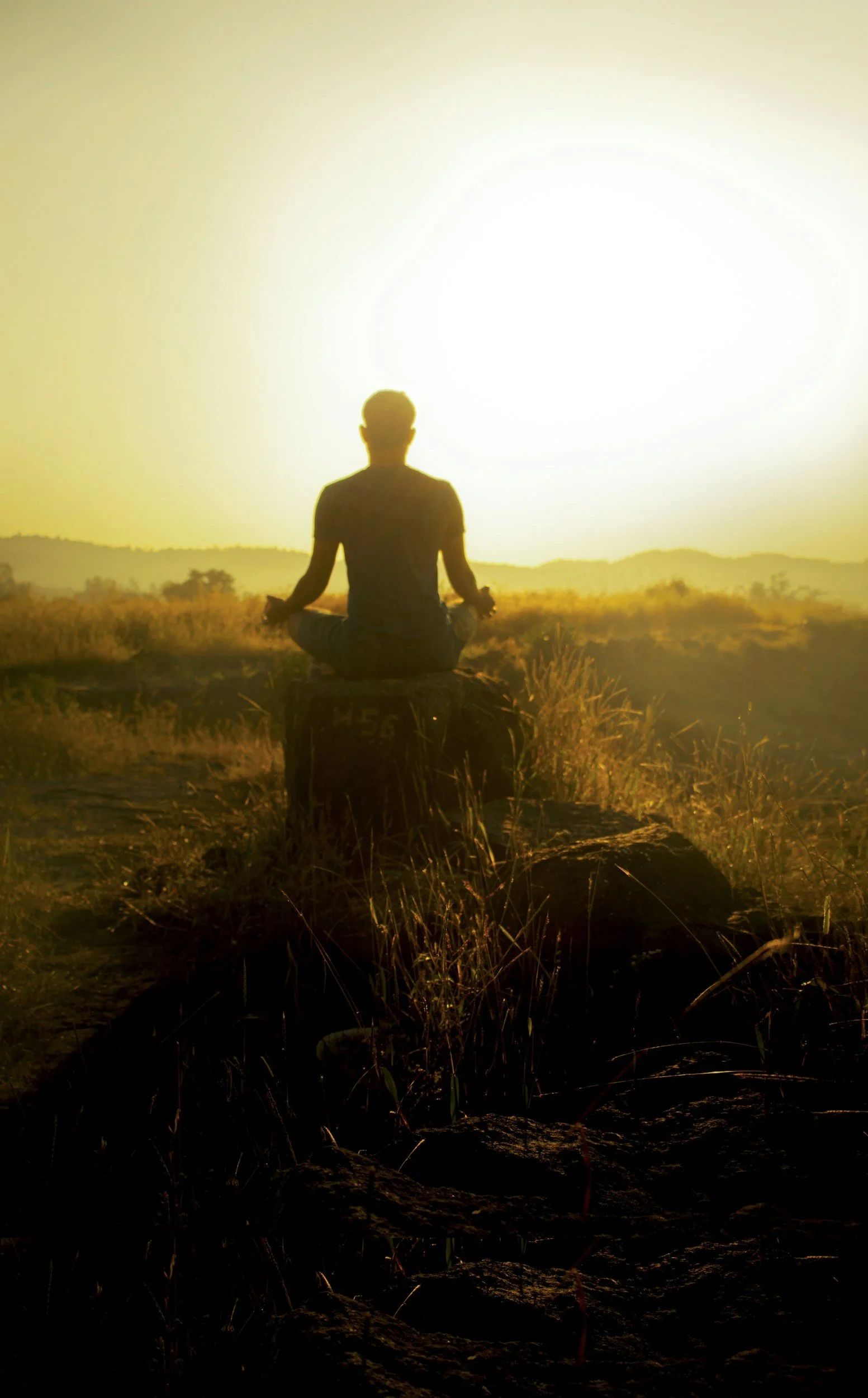 Silhouette of a person in a seated meditation pose on a rock at sunset or sunrise in an open field with hills in the background.