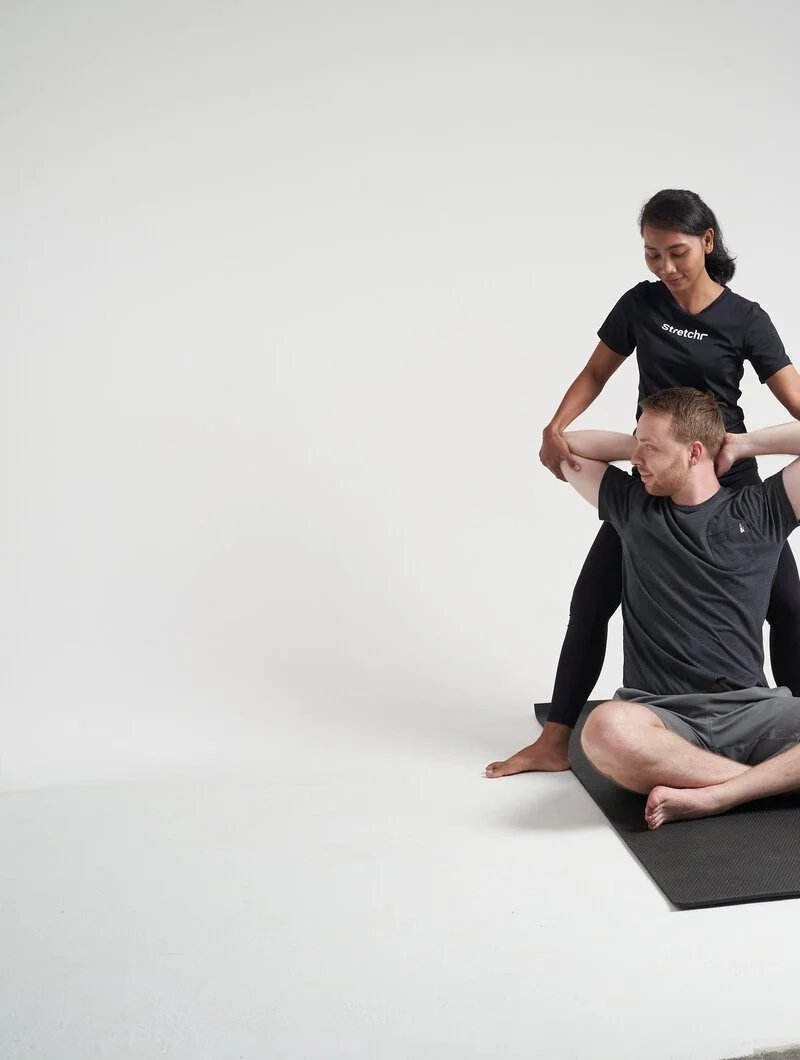 A woman is standing behind a man seated on the floor while holding onto his arms.