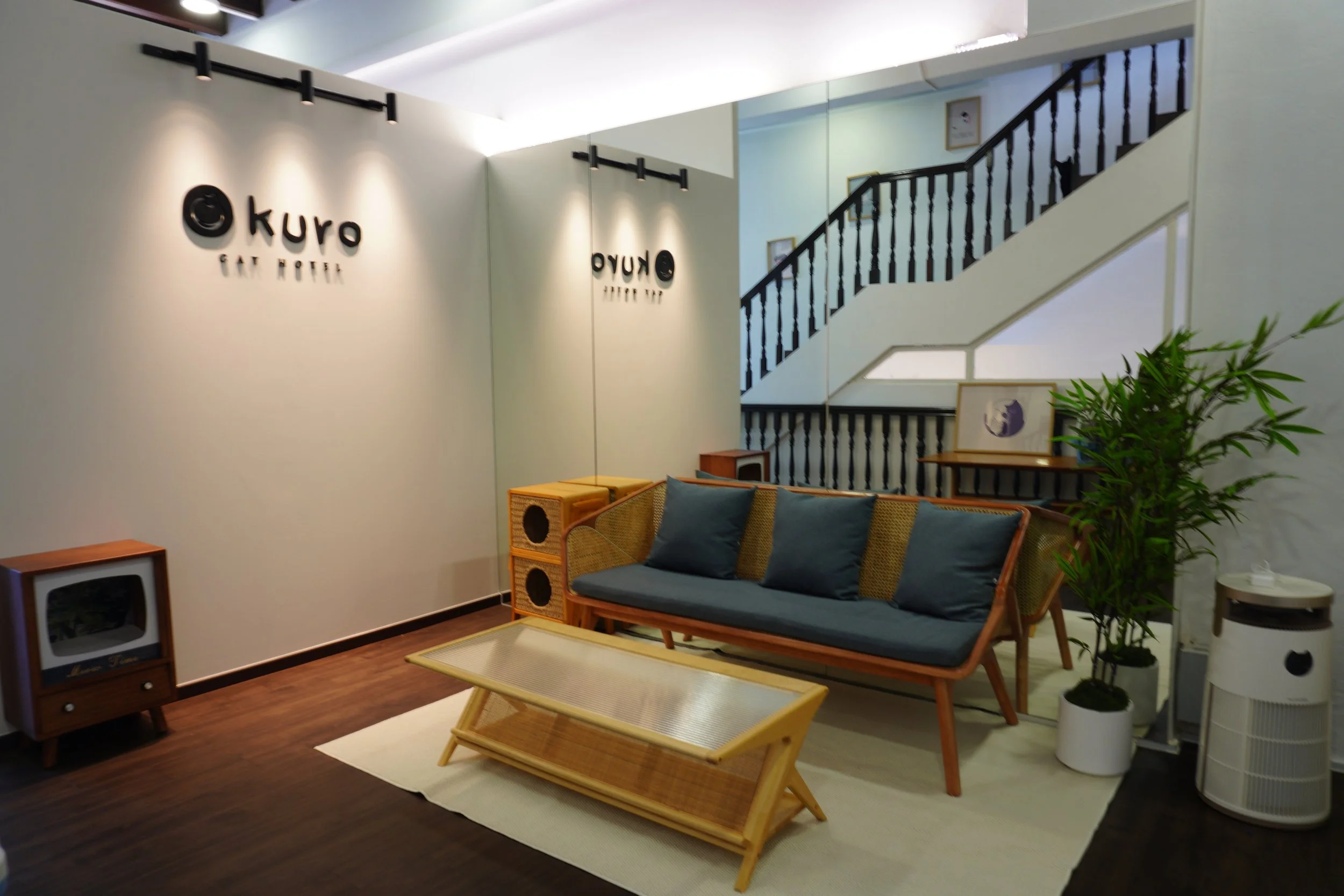 Interior of Kuro Say Hotel lobby featuring a vintage TV, a wooden coffee table, a navy blue sofa with three cushions, potted plants, a staircase with black railing, framed artwork on the wall, and a white air purifier.