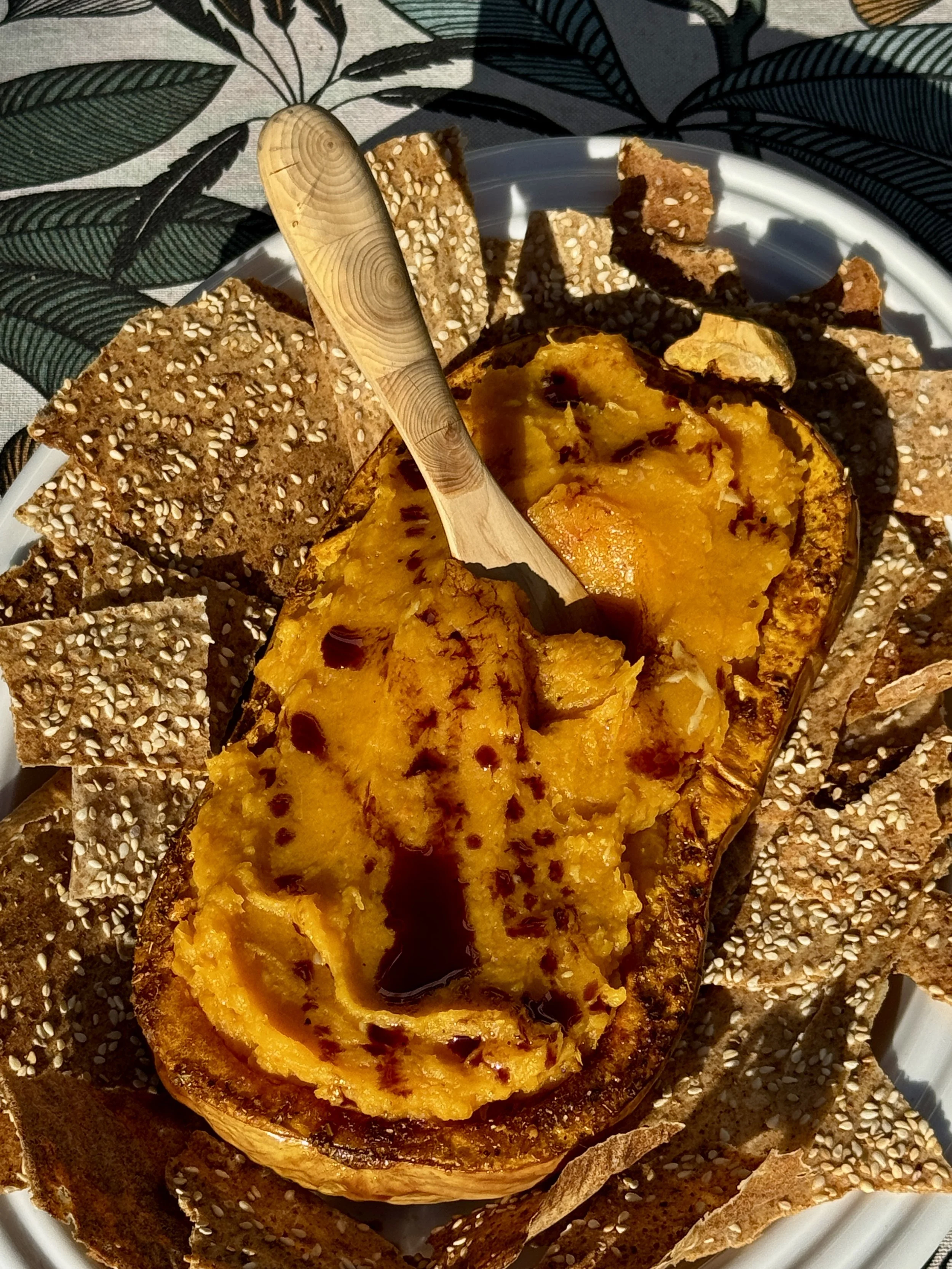 Baked and mashed butternut squash served in its skin topped with pumpkin seed oil, surrounded by sourdough crackers on a white plate.