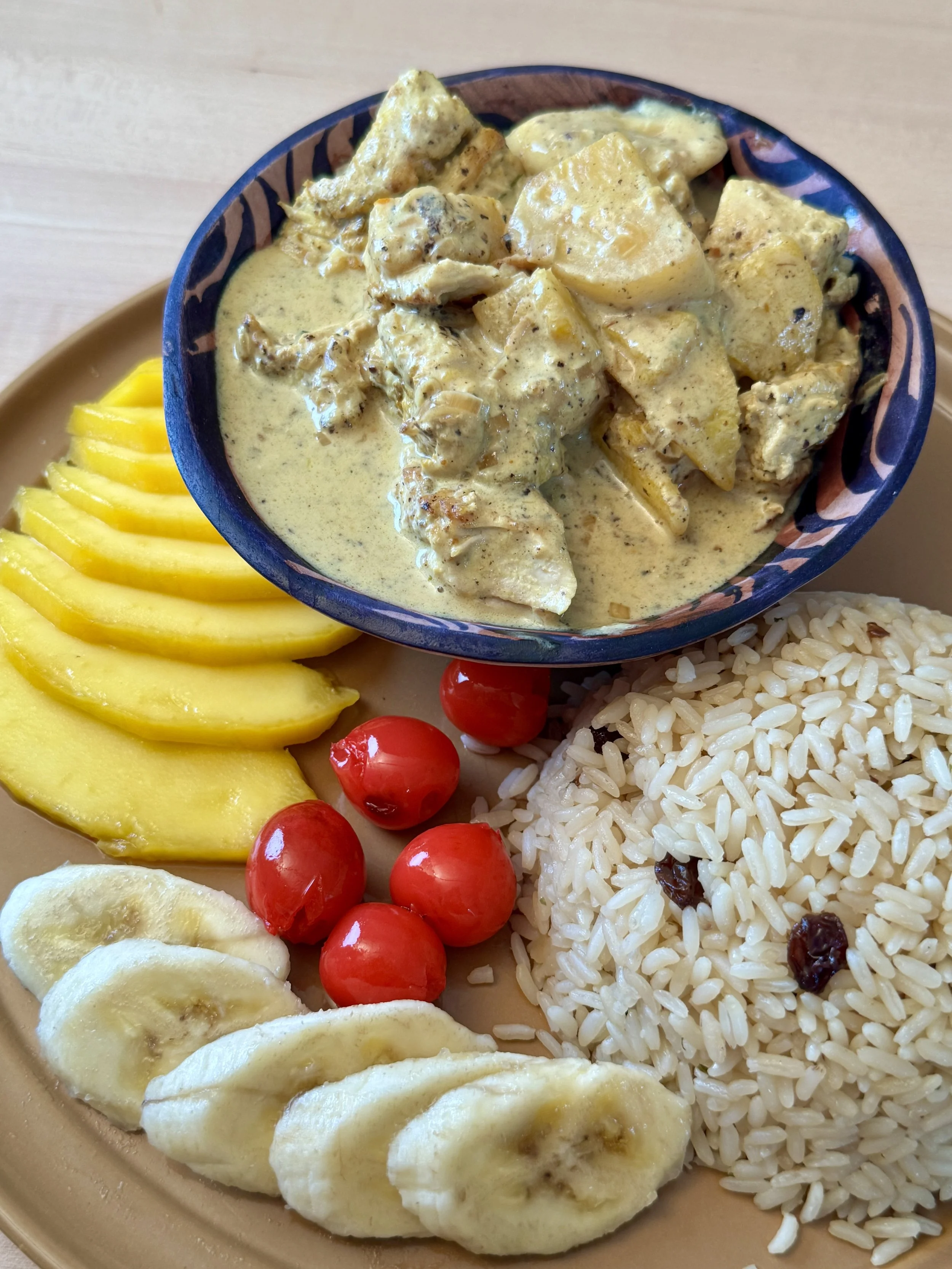 A plate of food originating in Switzerland called Riz Casimir, including sliced bananas, canned cheries, yellow mango slices, seasoned rice with raisins, and a bowl of chicken in creamy curry sauce mixed with pineapples.