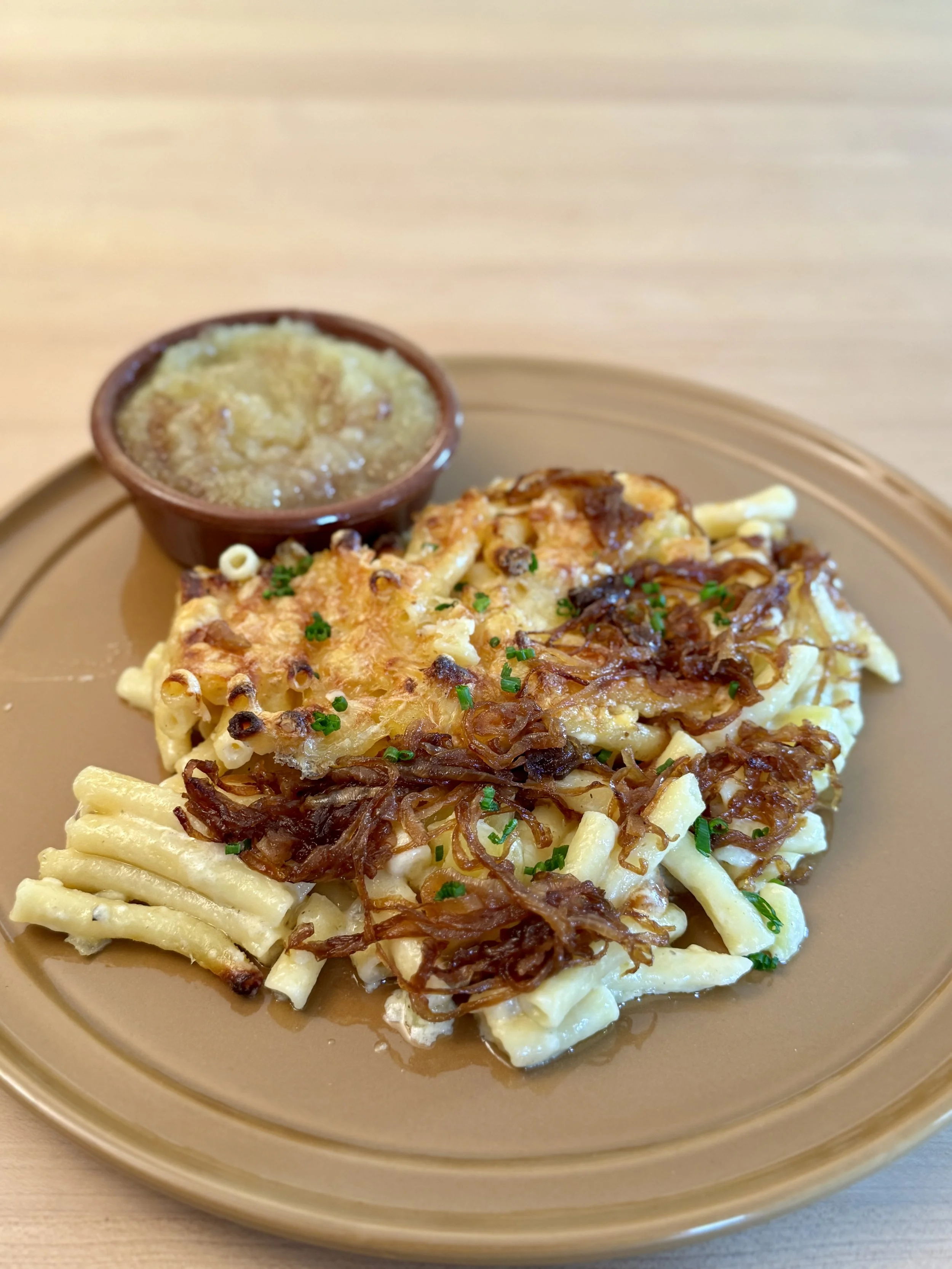 Baked Älplermagronen (similar to macaroni and cheese) topped with crispy fried onions, garnished with chopped green herbs, served with a small bowl of apple sauce.