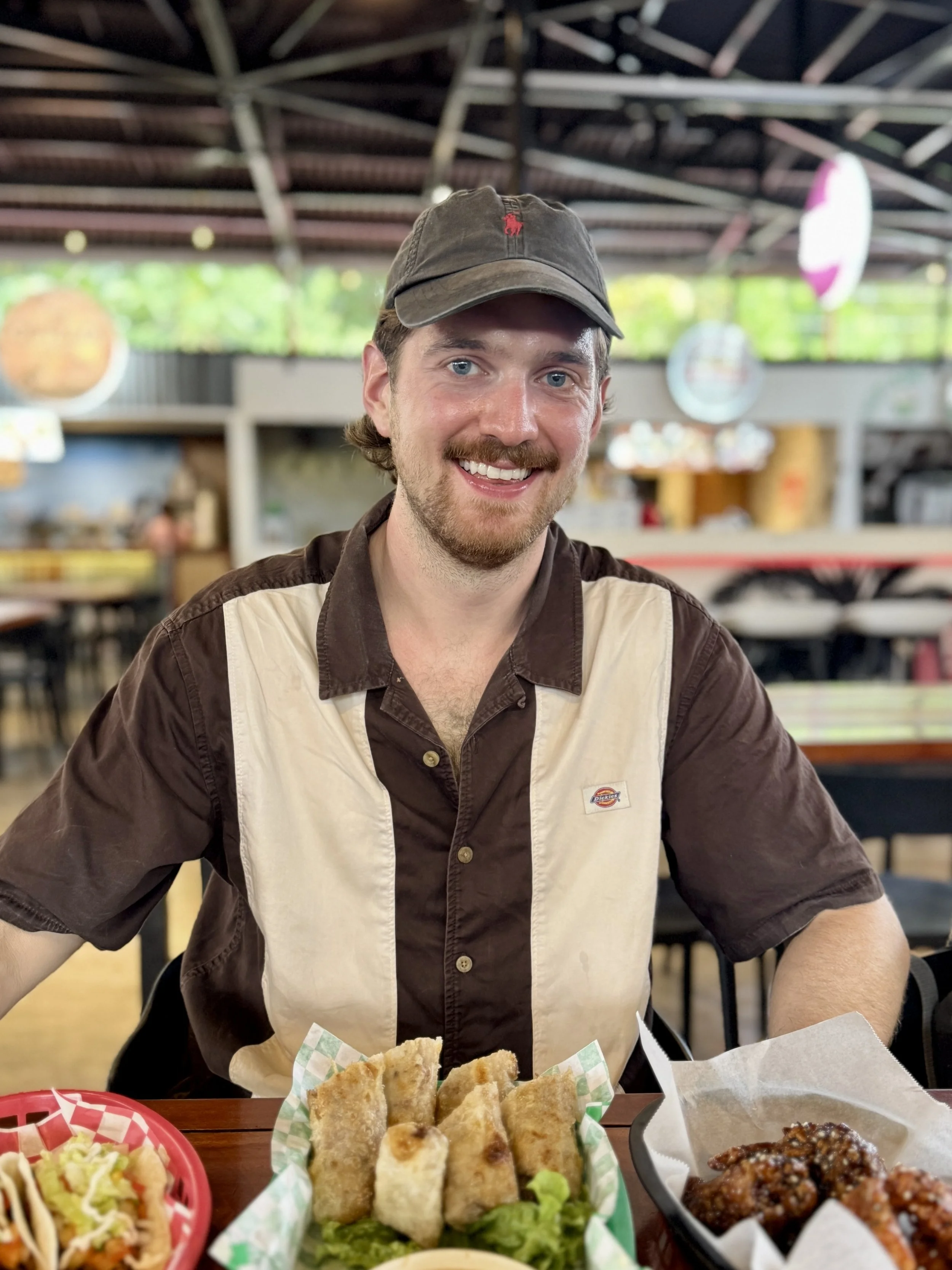 A smiling man with a beard and blue eyes wearing a brown and beige shirt and a gray cap sitting at a table with various Chinese food dishes, including spring rolls and fried chicken.