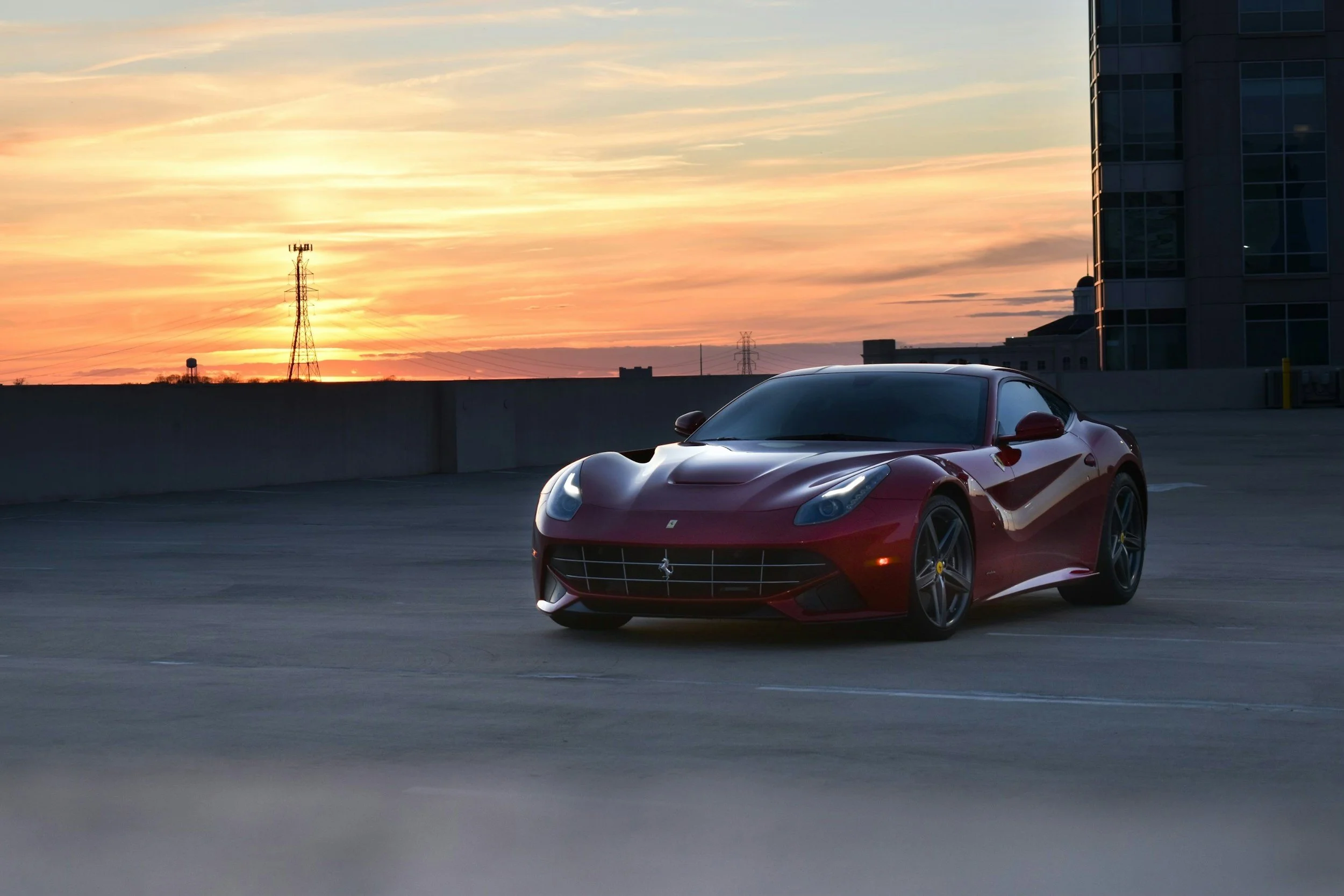 A red Ferrari sports car parked on the top level of a parking garage during sunset, with a cityscape and a sunset sky in the background.
