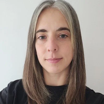 A woman with straight, shoulder-length brown hair and fair skin, wearing a black top, standing against a white background.