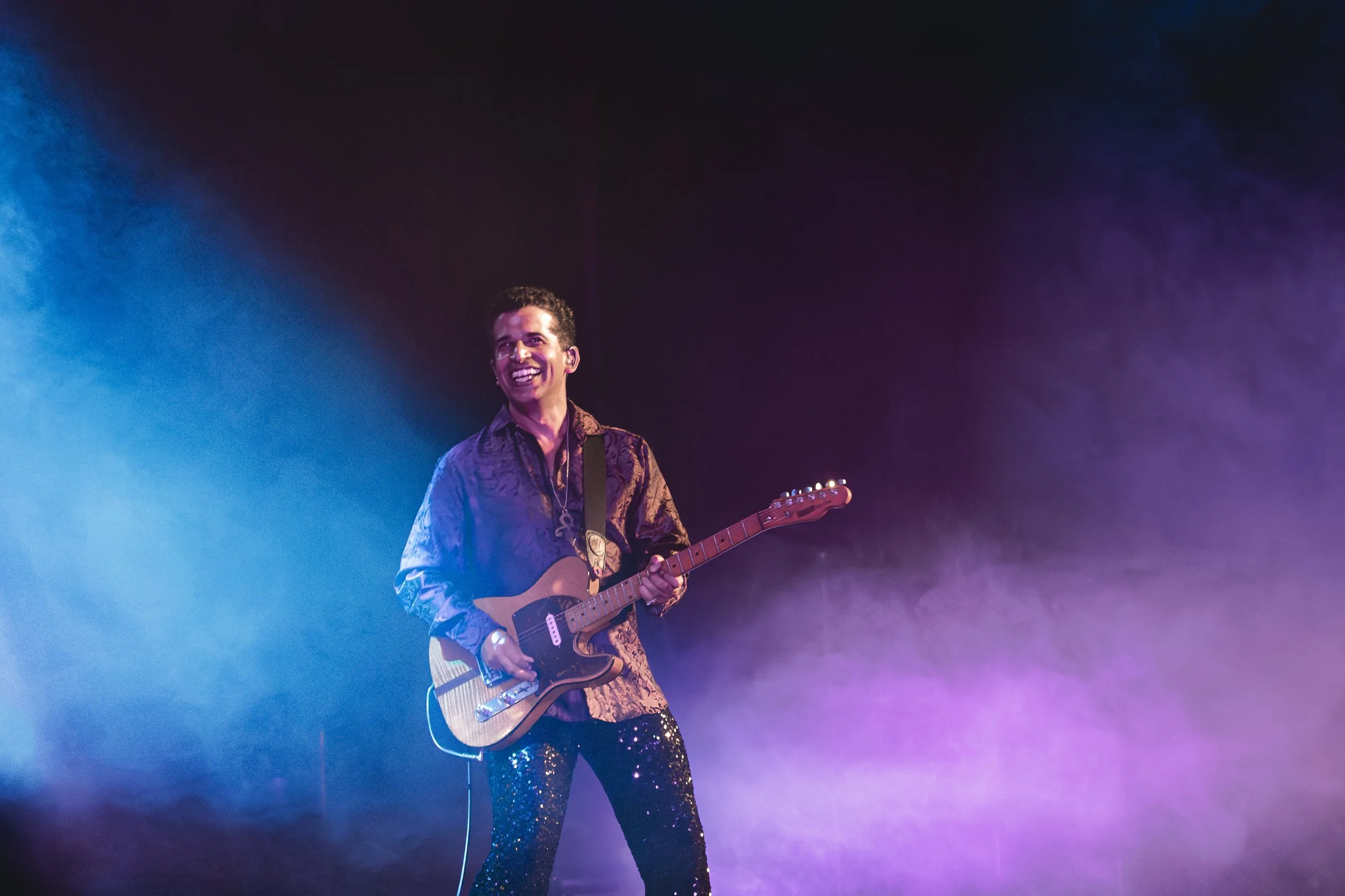 Prince tribute act, New Purple Celebration performing on stage with colourful lighting, playing an electric guitar, wearing a shiny toned outfit and smiling.
