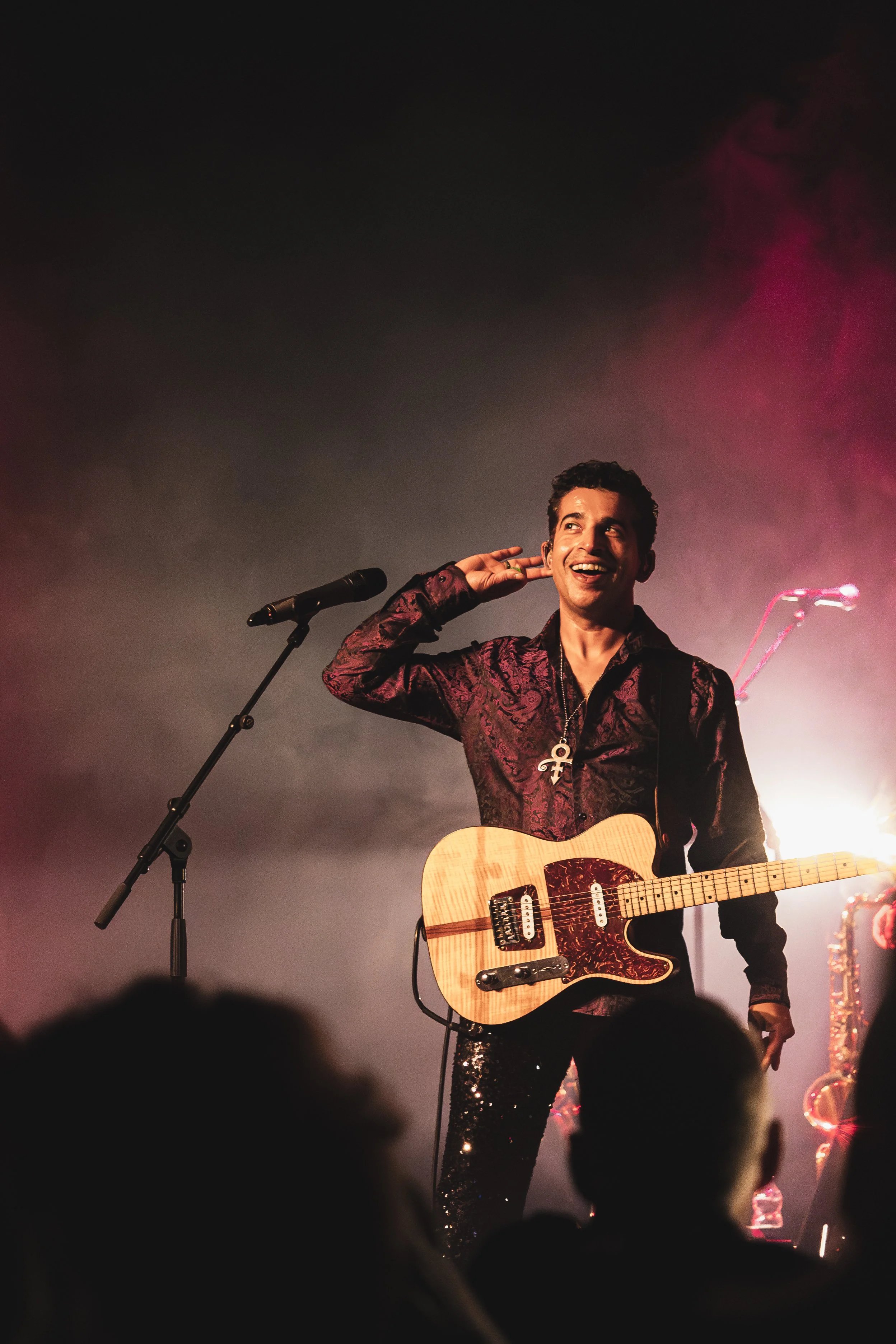 Prince tribute act on stage holding an acoustic electric guitar, smiling and wearing a dark patterned shirt with a cross necklace. Behind him, purple lighting and stage smoke create a vibrant atmosphere, with an audience silhouette in the foreground.