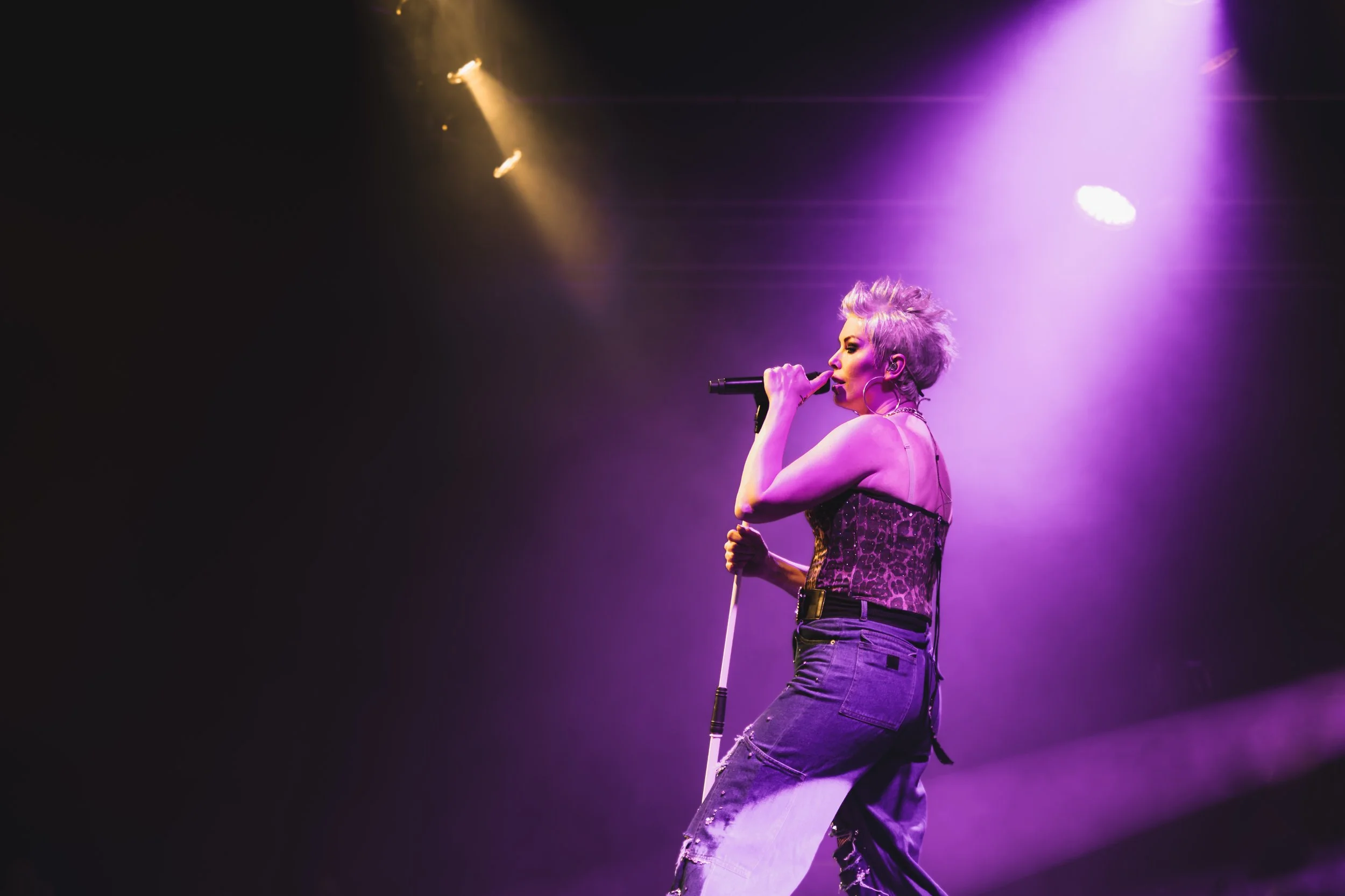 A female singer with short, purple hair performing on stage under purple and yellow spotlight lighting, holding a microphone.