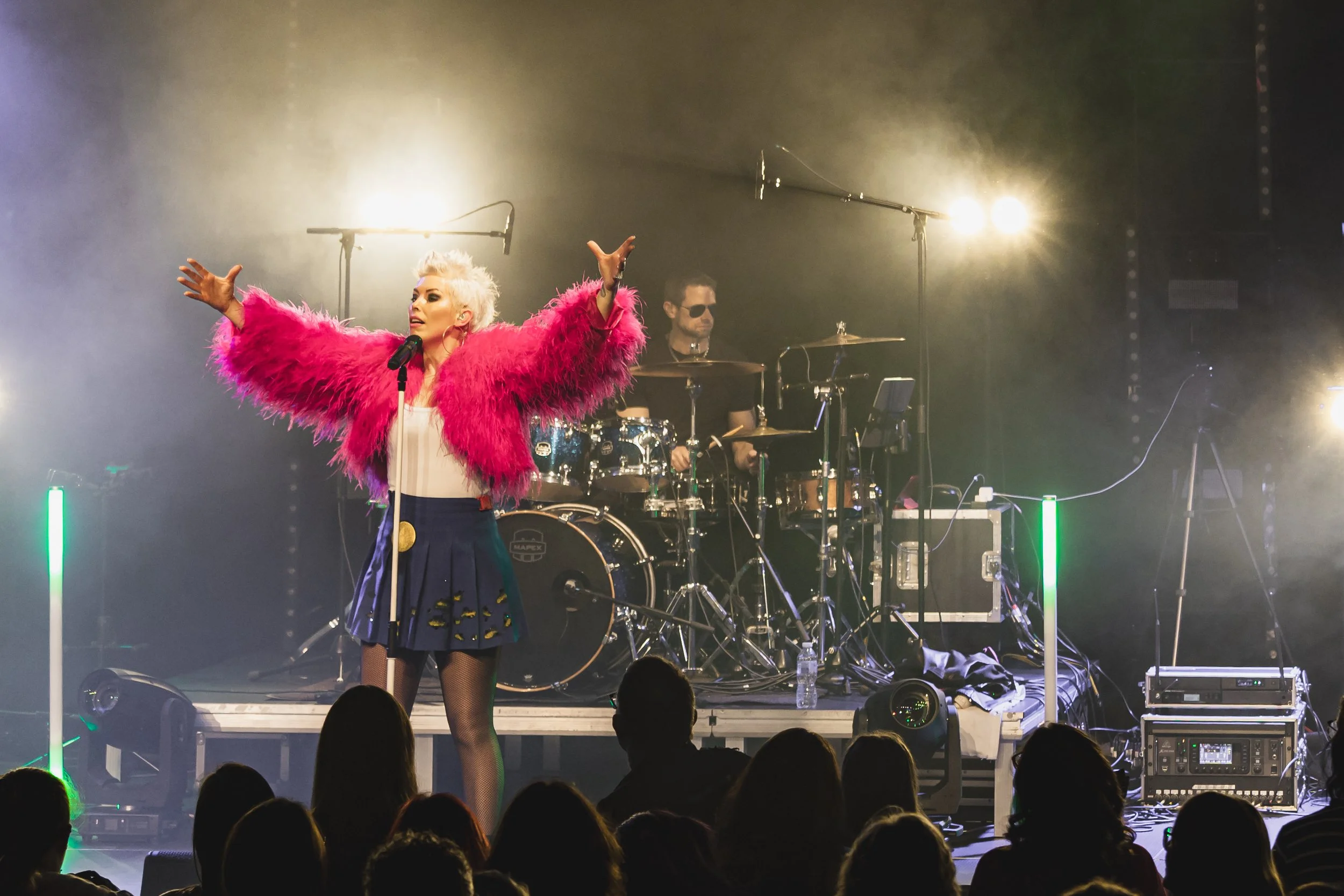 A female singer performing on stage with a band, wearing a bright pink fluffy jacket, a white top, a dark skirt, and fishnet stockings, with a drummer playing behind her and an audience in front.