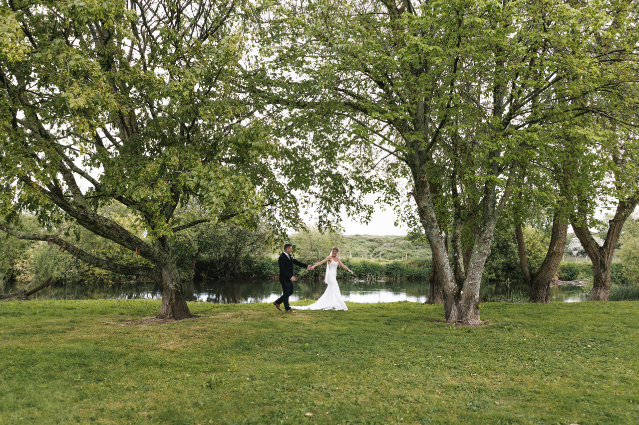 Bride and groom walking by the river  on their wedding day at the Bridge House Hotel in Ferndown, Dorset.