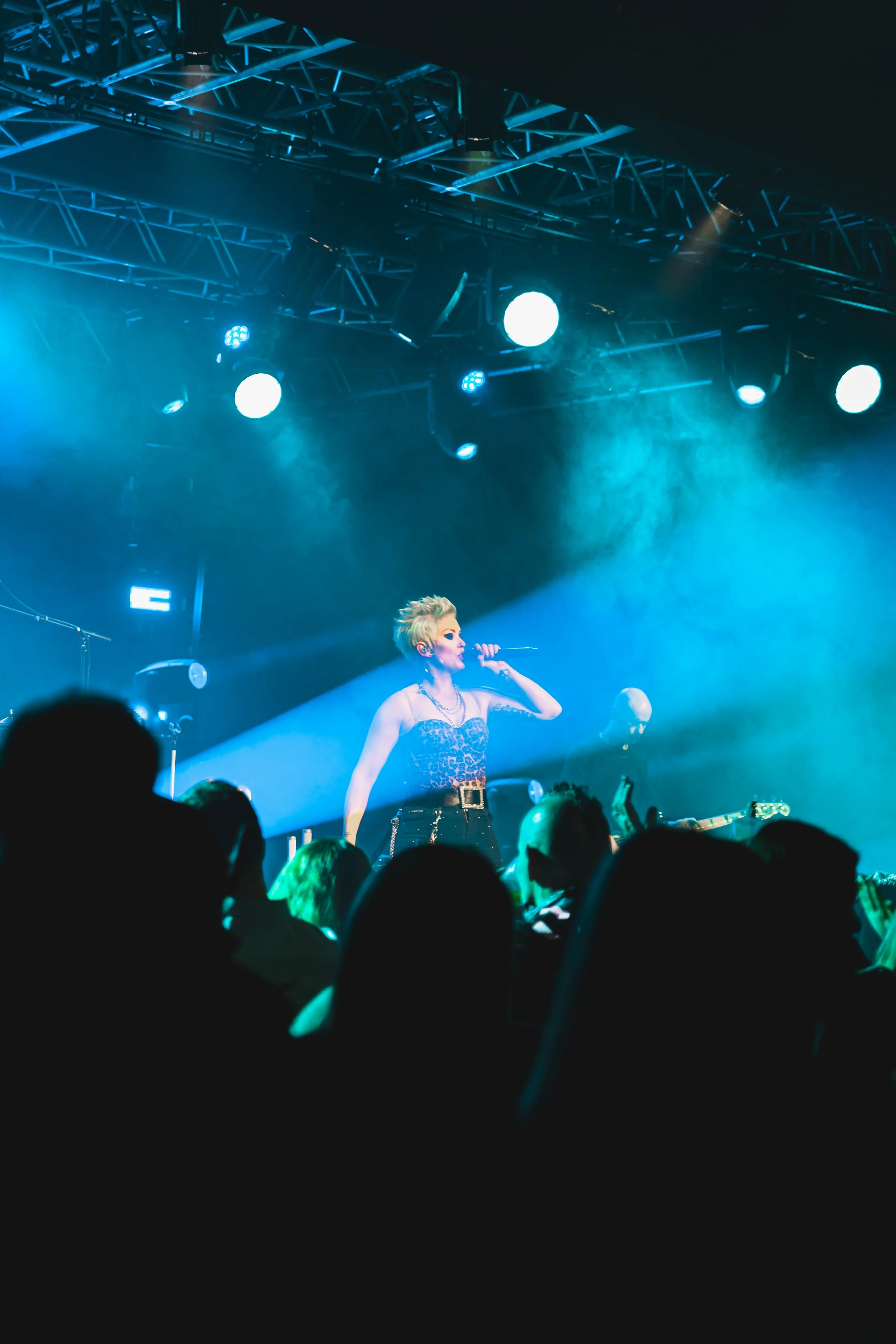 A female singer with short, spiky hair performing on stage with blue lighting, wearing a leopard print top and black pants, facing an audience.
