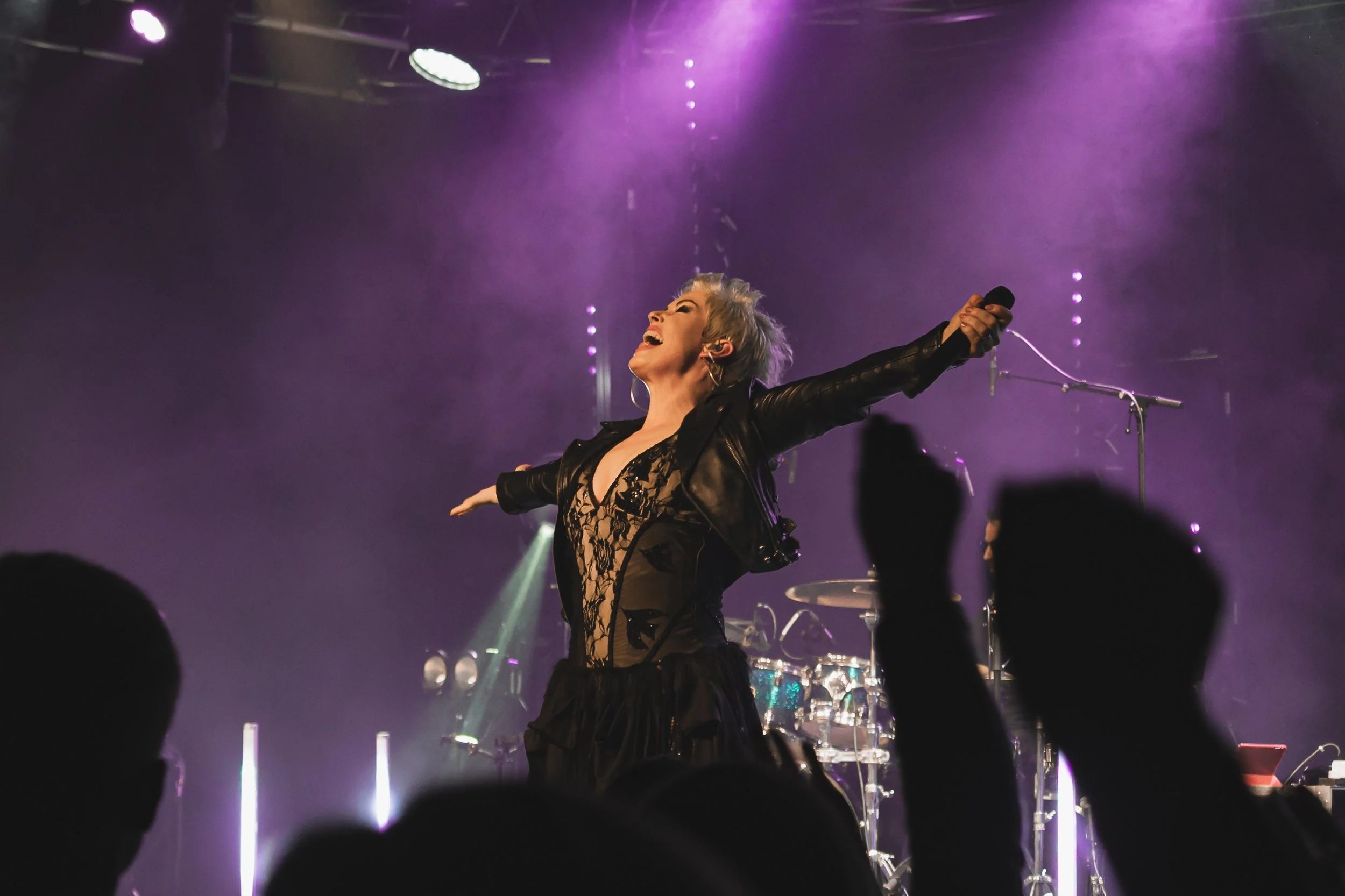 Performer on stage with arms outstretched, singing or smiling, wearing a black lace dress and leather jacket, with purple stage lighting and an audience in the foreground.