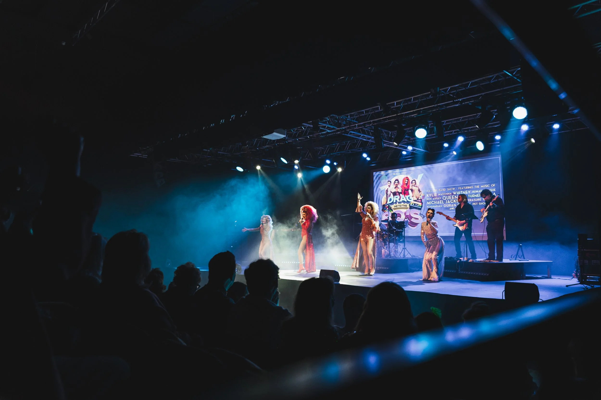 A live performance on stage with singers in colorful costumes and band members playing guitars, with an audience watching in a dark concert hall.