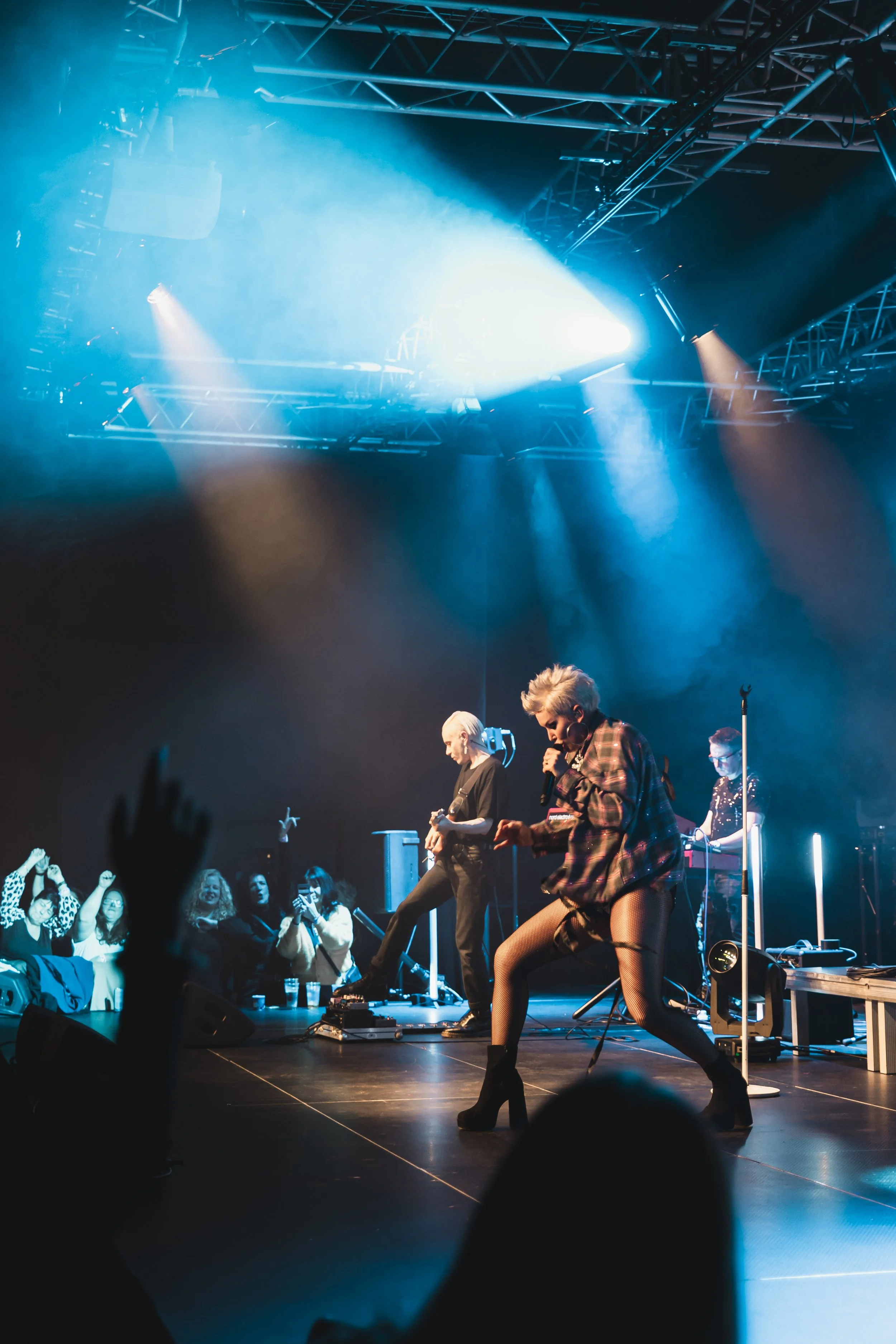 A live concert with a female singer performing on stage, dancing energetically, accompanied by band members with instruments, under colorful stage lighting, with an engaged audience.