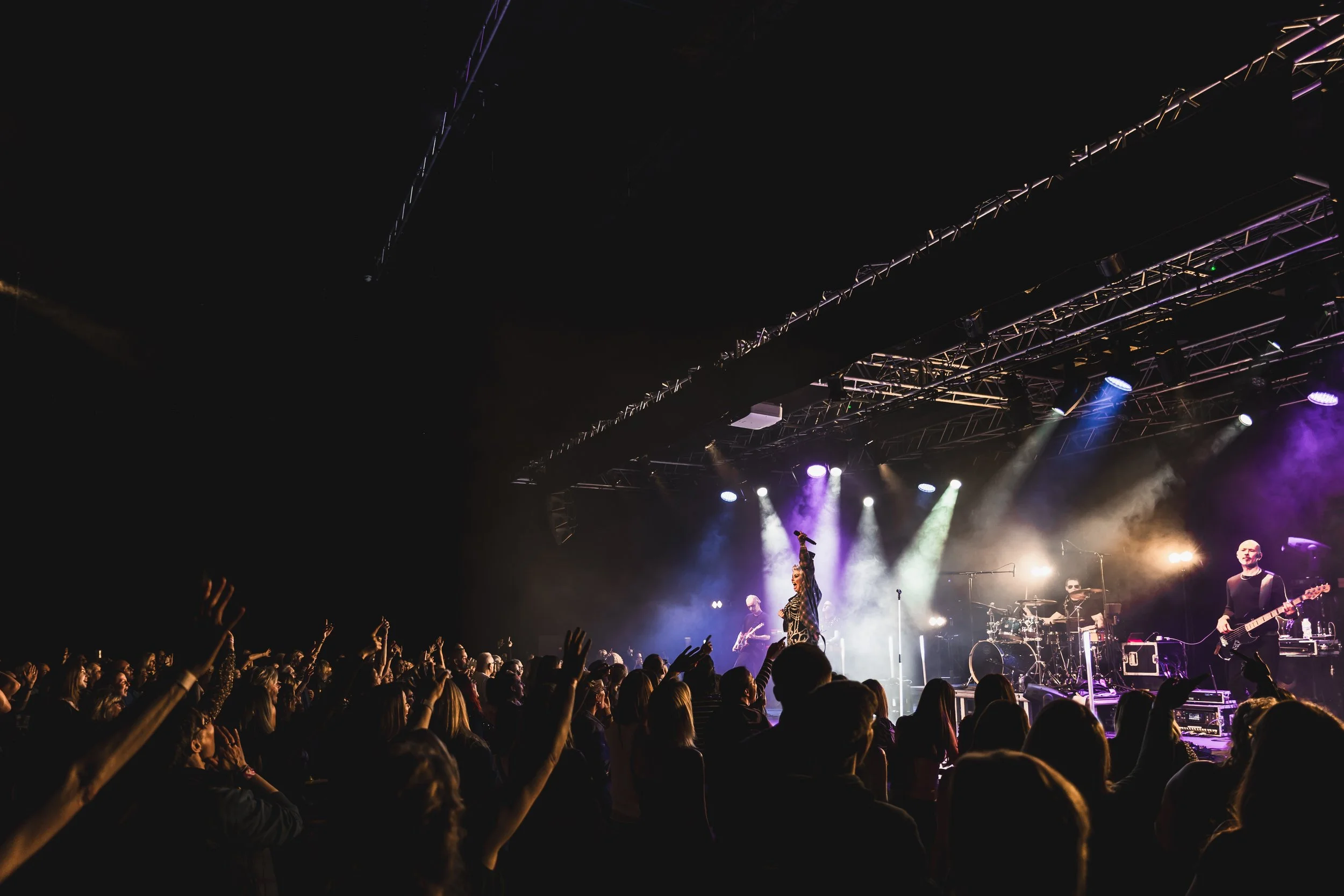 Concert scene with band performing on stage under colorful lights and an audience with raised hands.