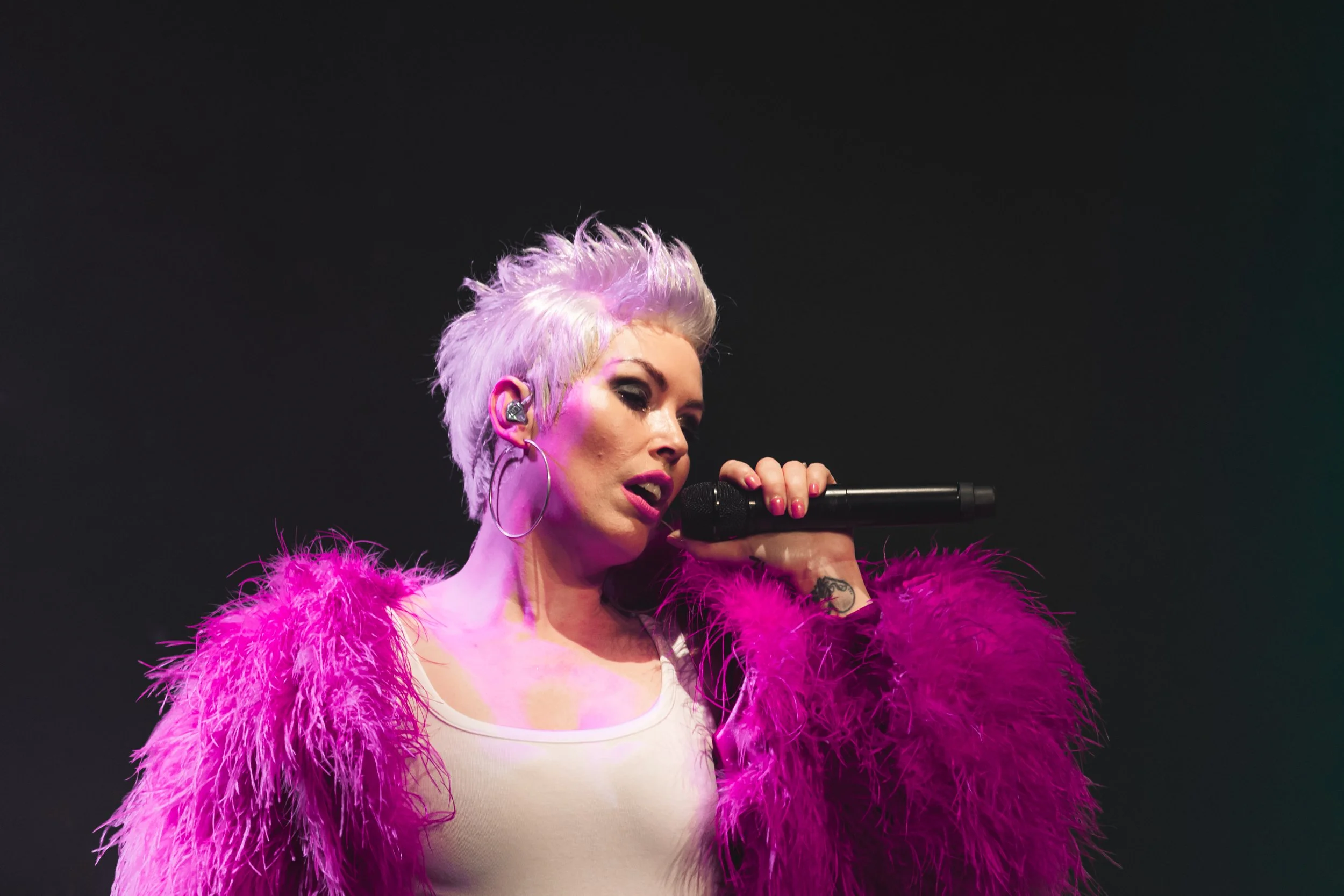 A female singer with short, spiky lavender hair singing into a microphone on stage, wearing a pink feathered jacket and large hoop earrings.