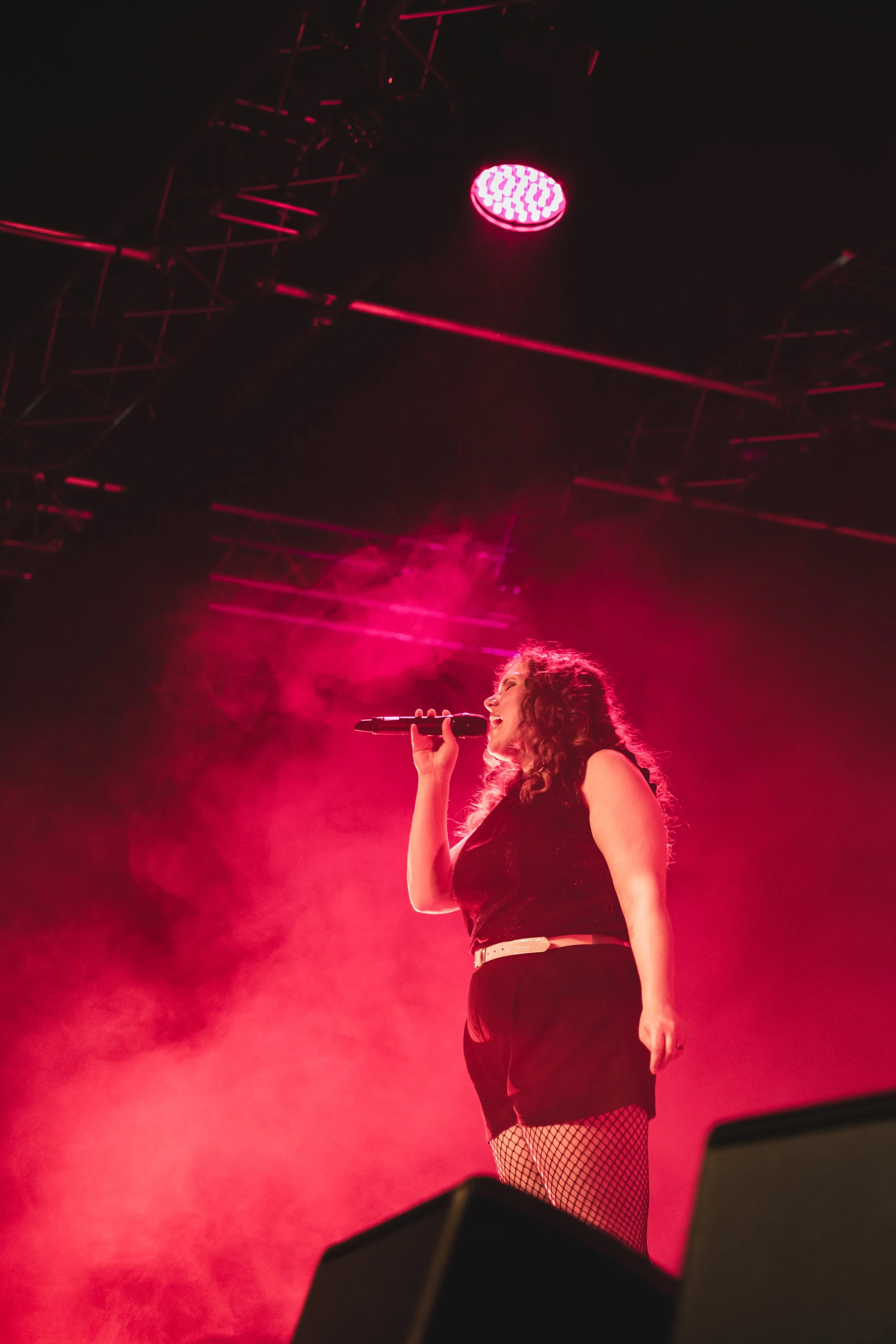 A woman singing on stage with pink lighting and fog, wearing a black outfit and fishnet stockings.