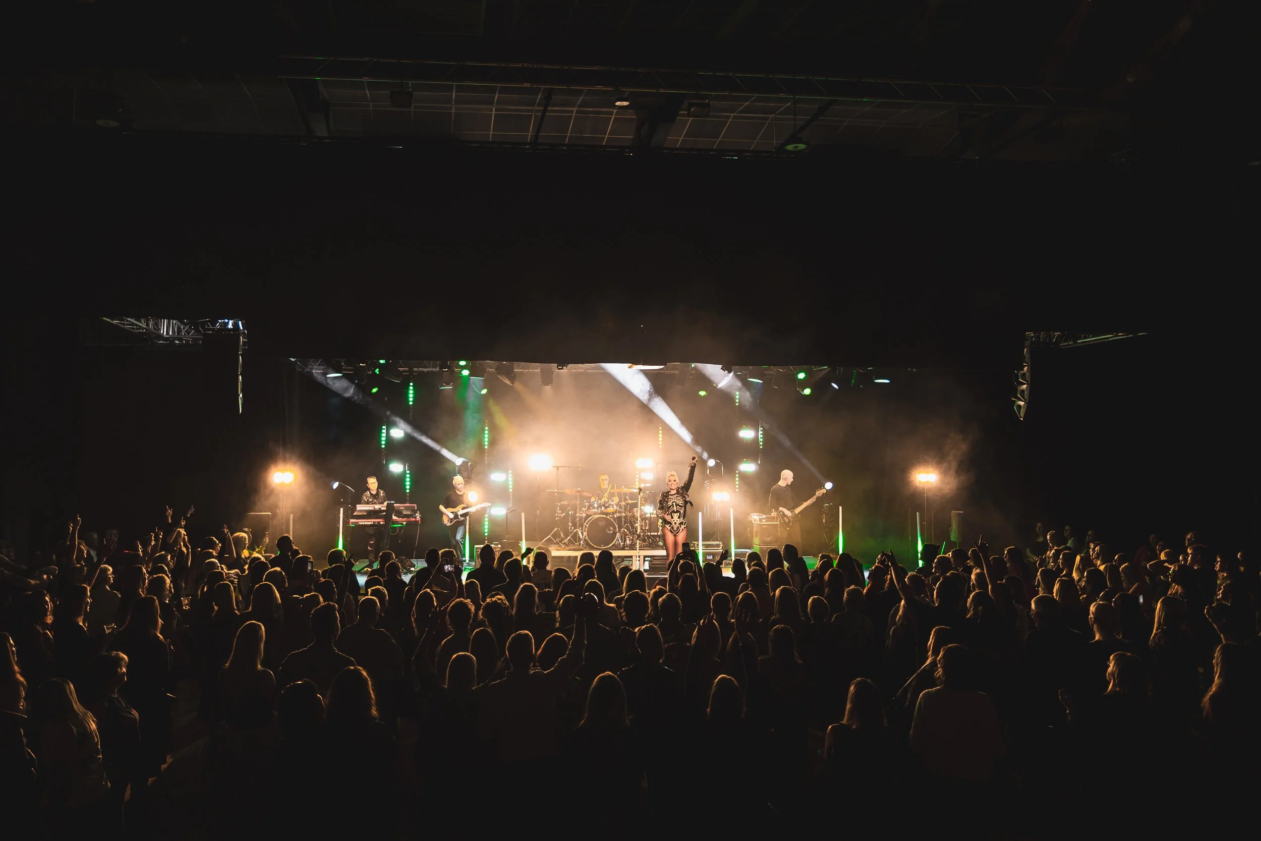 A concert performance on stage with band members playing instruments and singer with arm raised, illuminated by bright lights, with an audience in the foreground.